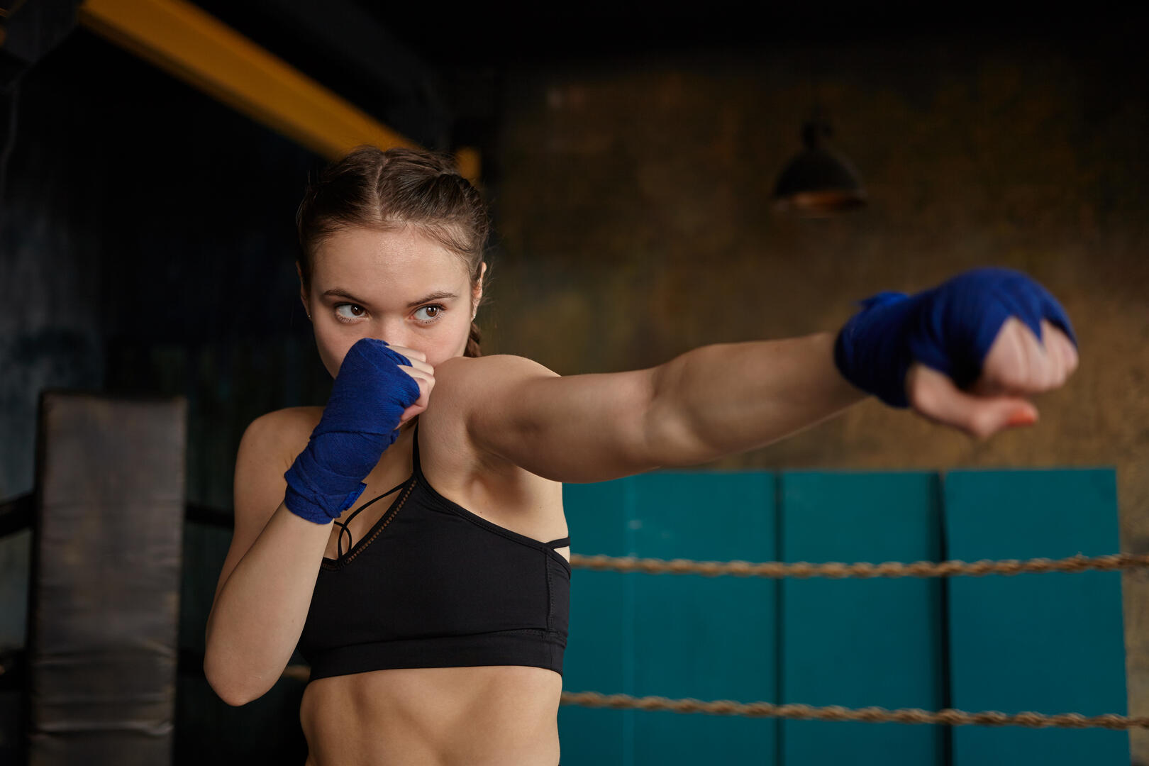 Cropped portrait of self determined young woman professional boxer with strong muscular arms and abdominal wearing black sports top and blue boxing bandages mastering punching technique in gym