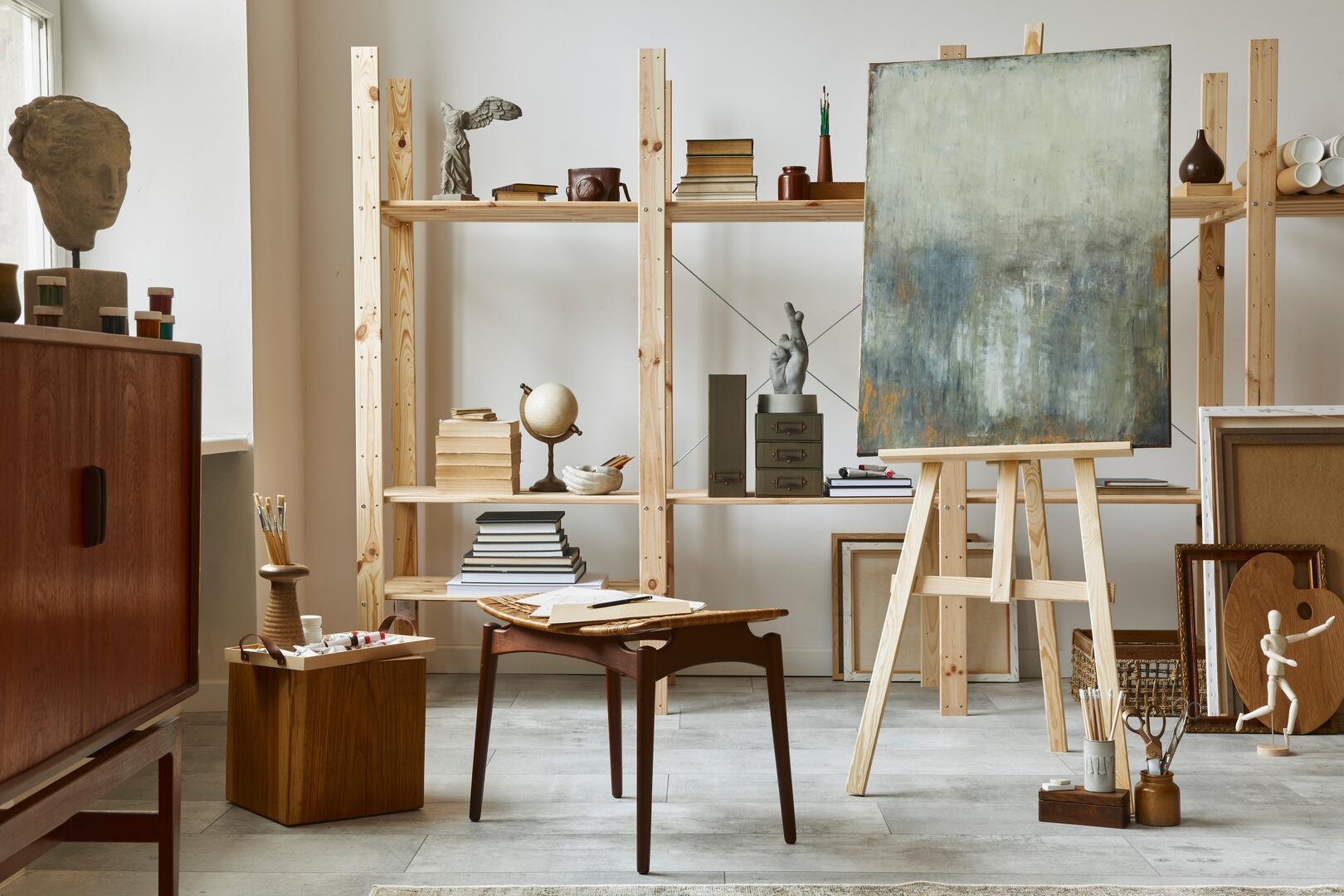 Unique artist workspace interior with stylish teak commode, wooden easel, bookcase, artworks, painting accessories, decoration and elegant personal stuff. Modern work room for artist. Template.