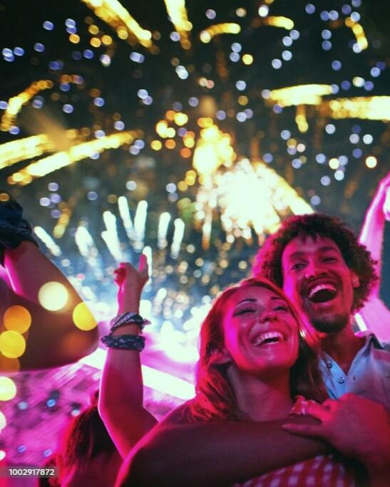 Closeup low angle view of group of multi ethnic young adults dancing and enjoying an open air concert on a summer afternoon. They are standing in a circle while fireworks popping in the background.