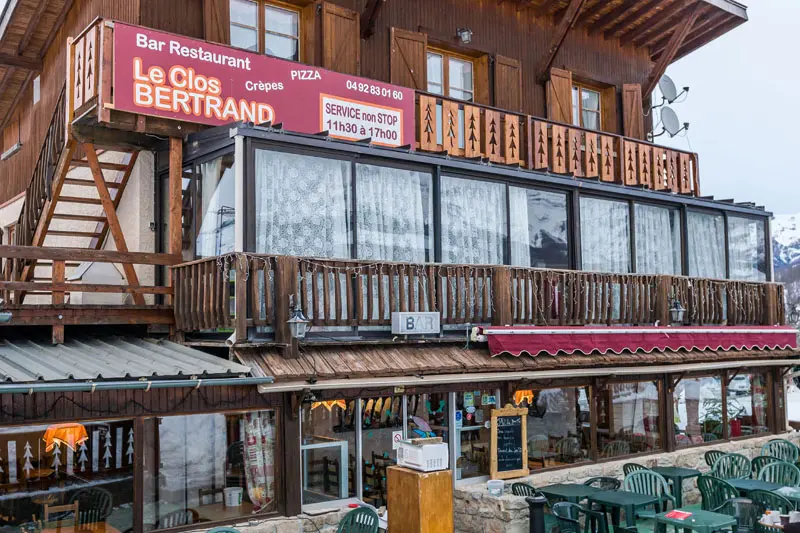 Restaurant with an outdoor terrace with tables and chairs, and a two-story stone main building. A sign on the second floor reading: 