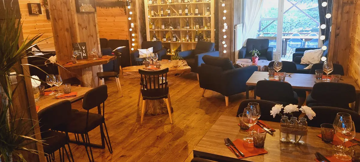 Intérieur du restaurant, ambiance chaleureuse avec fauteuils confortables, intérieur bois, tables en bois, bouteilles de vin en présentoir, porte-fenêtre donnant sur la terrasse de l'établissement
