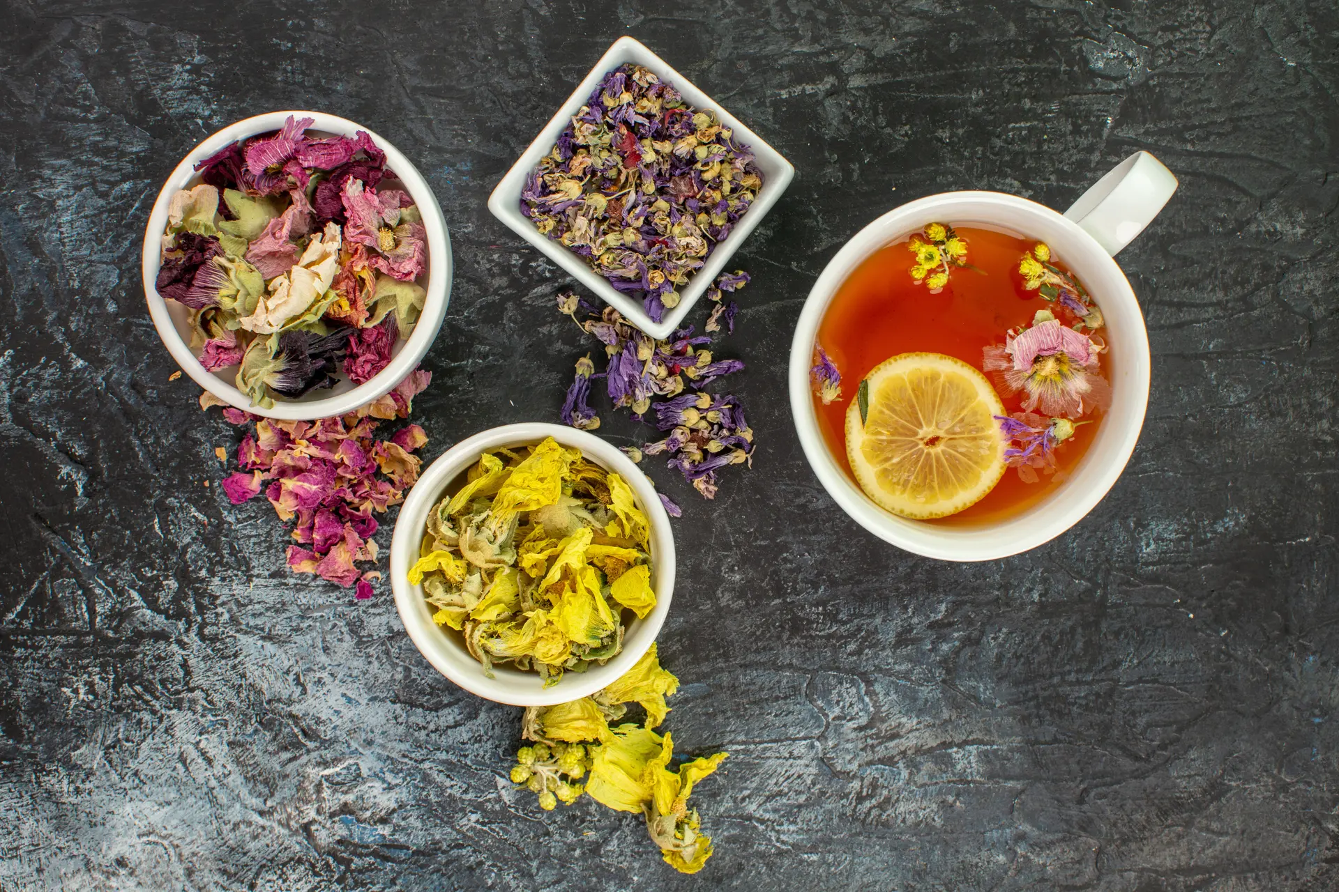 close up photo of herbal tea near bowls of dry flowers on grey background