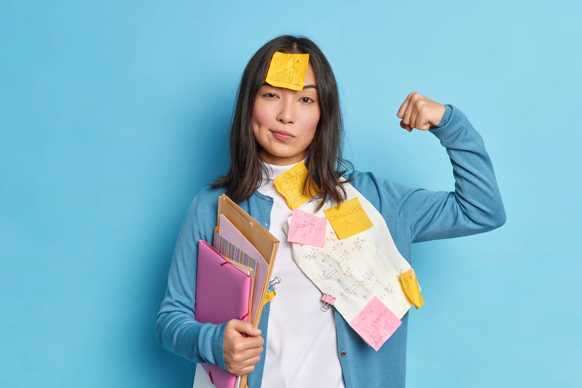 Powerful female student raises arm and shows muscles feels confident after working on diploma paper wears stickers on forehead holds folders isolated over isolated over blue studio background