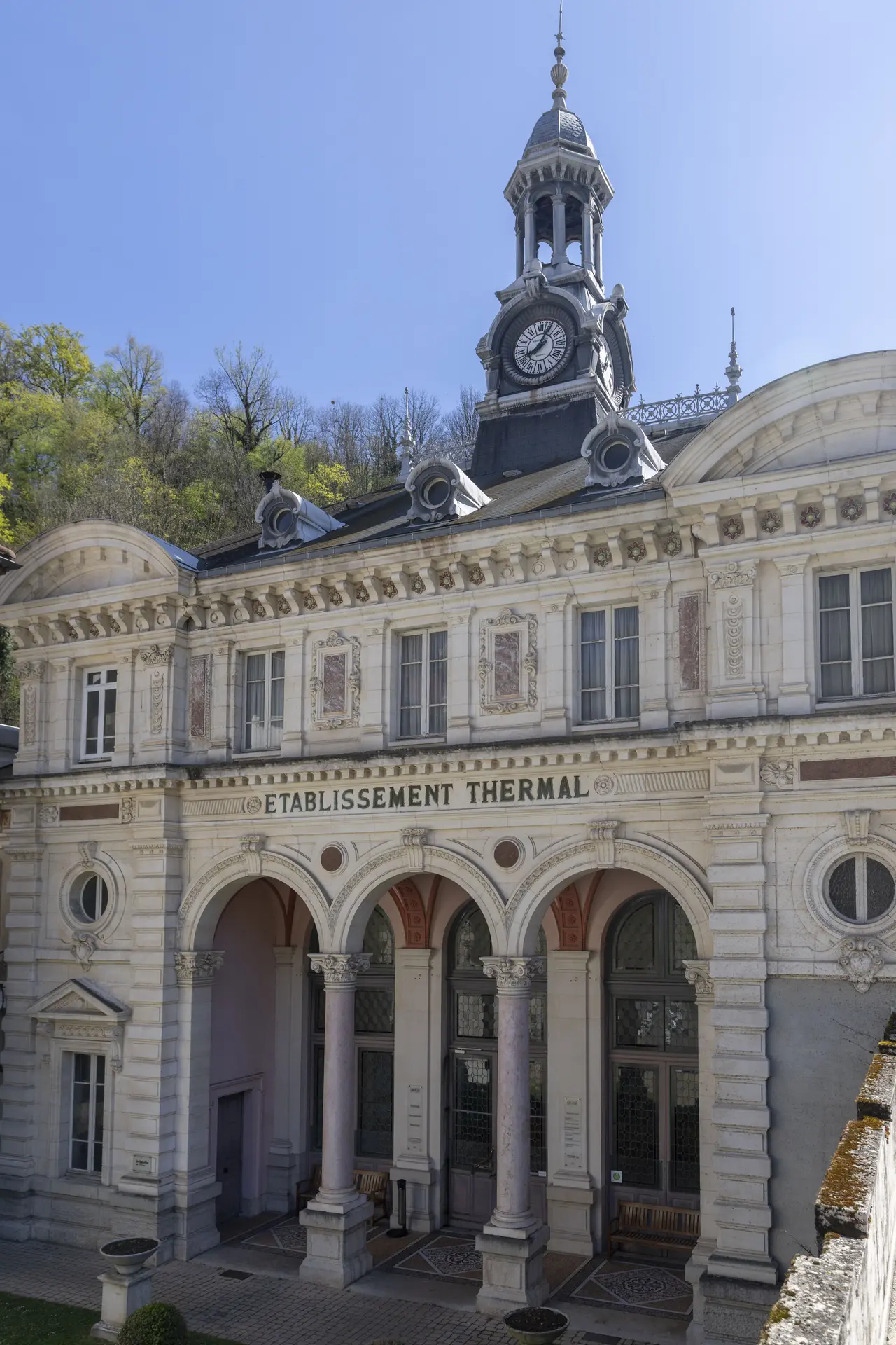 Le bâtiment des thermes d'Uriage a une façade symétrique de style classique, avec trois arches d'entrée surmontées de l'inscription ÉTABLISSEMENT THERMAL, une horloge dans une tourelle coiffée d'une flèche, et des murs ornés de fenêtres rectangulaires et de médaillons.