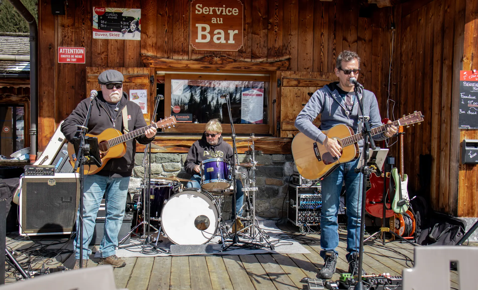 Three musicians play on a wooden terrace in front of a mountain bar. The band consists of two guitarists/singers and a drummer in the center.