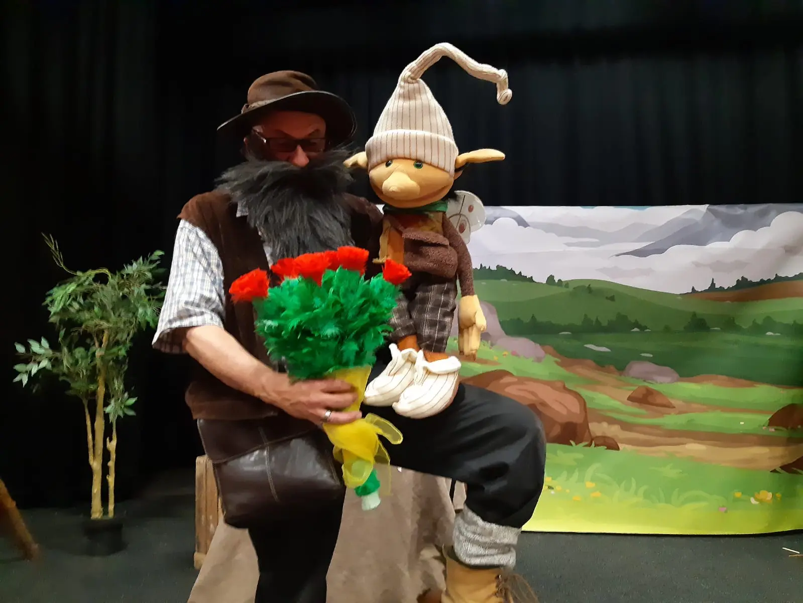 Yann, comédien, assis sur scène, déguisé en lutin avec une marionnette dans une main et un bouquet de roses rouges dans l'autre.
