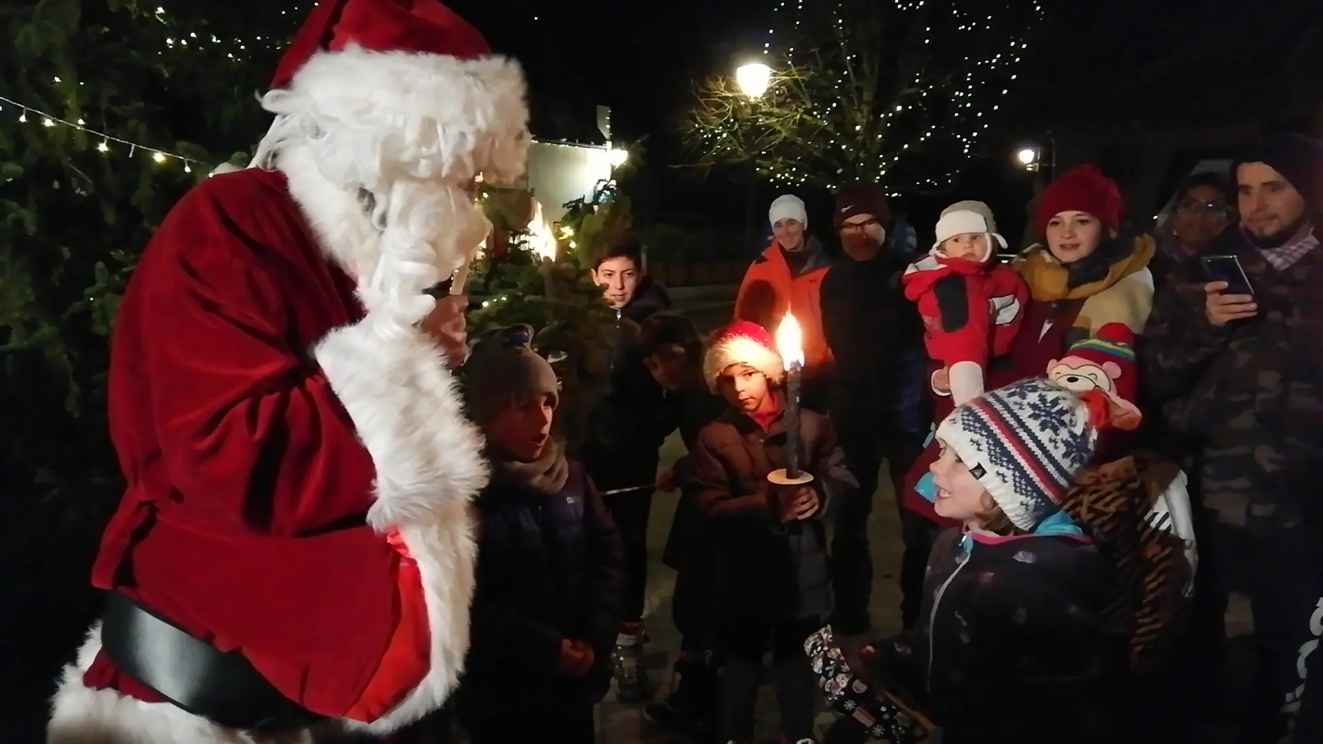 Le Père-Noël distribue les cadeaux aux enfants, à la lumière des flambeaux, lampions et guirlandes lumineuses.