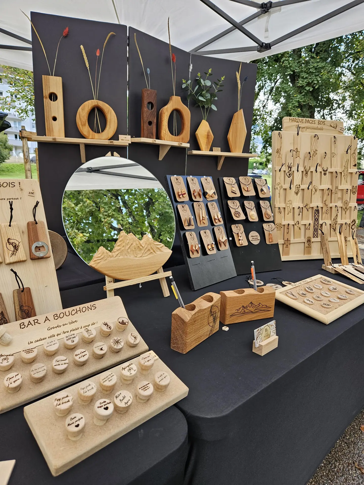 Ce stand présente une variété d'objets en bois, incluant des vases, des bijoux, des marque-pages et des bouchons gravés, disposés sur une table noire avec un miroir central.