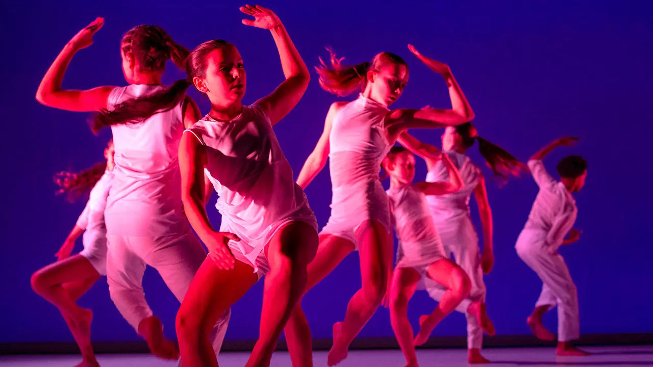 7 jeunes danseurs habillés sur blanc bougent sur scène dans un éclairage rose et bleu violet