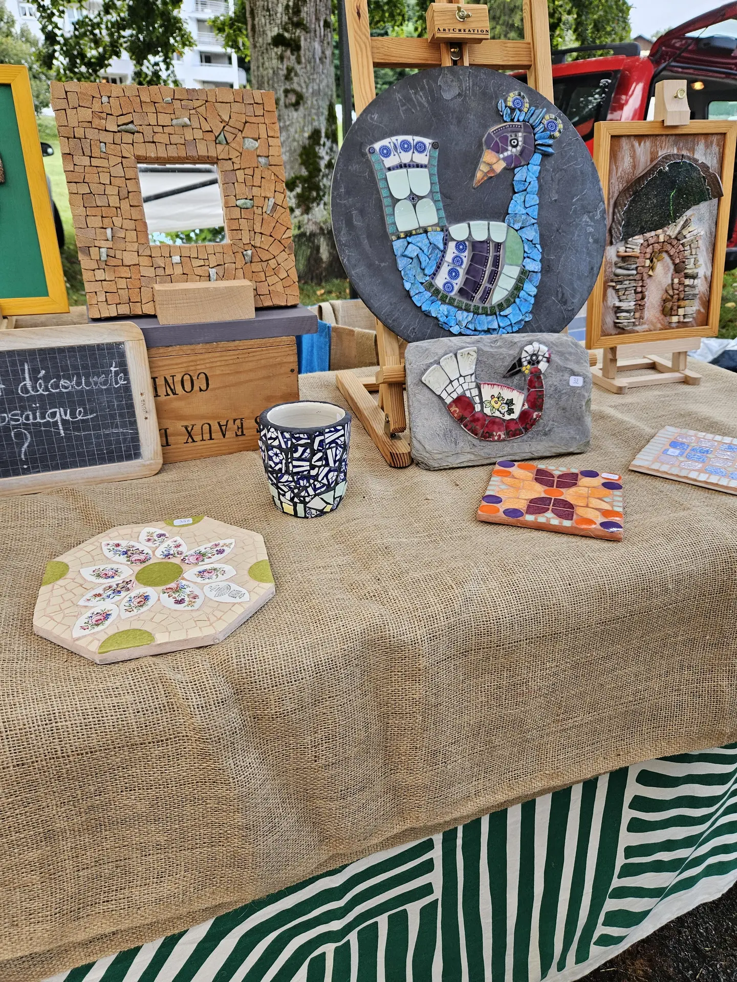 Ce stand de marché des créateurs présente une variété d'objets artisanaux, incluant des cadres, des articles en bois et d'autres créations, disposés sur une table recouverte d'un tissu beige avec des motifs verts et blancs rayés.