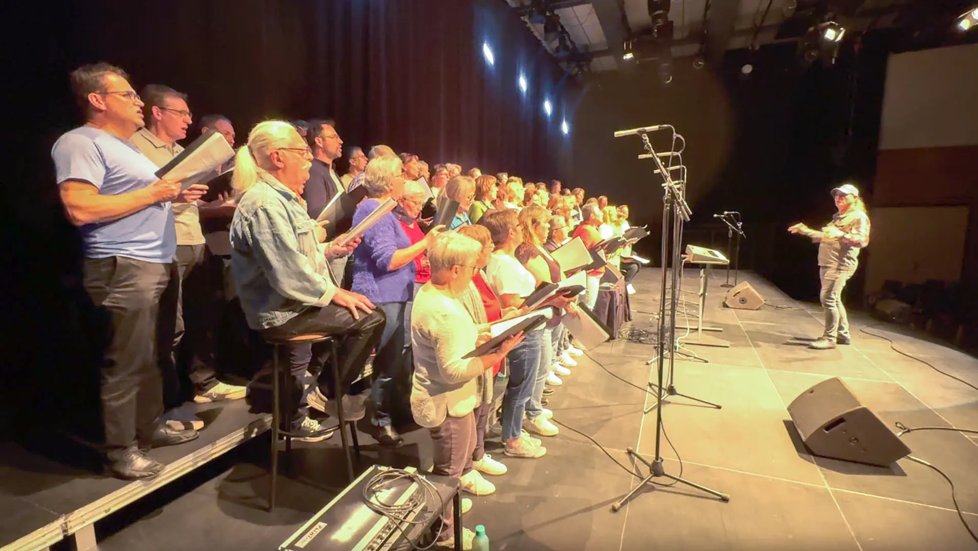 Photo d'un groupe de personnes sur scène. Le groupe chantent avec des partitions dans les mains. Sur le devant de la scène, le chef de choeur, dirige le groupe. Des micros sur pieds sont placés devant le groupe