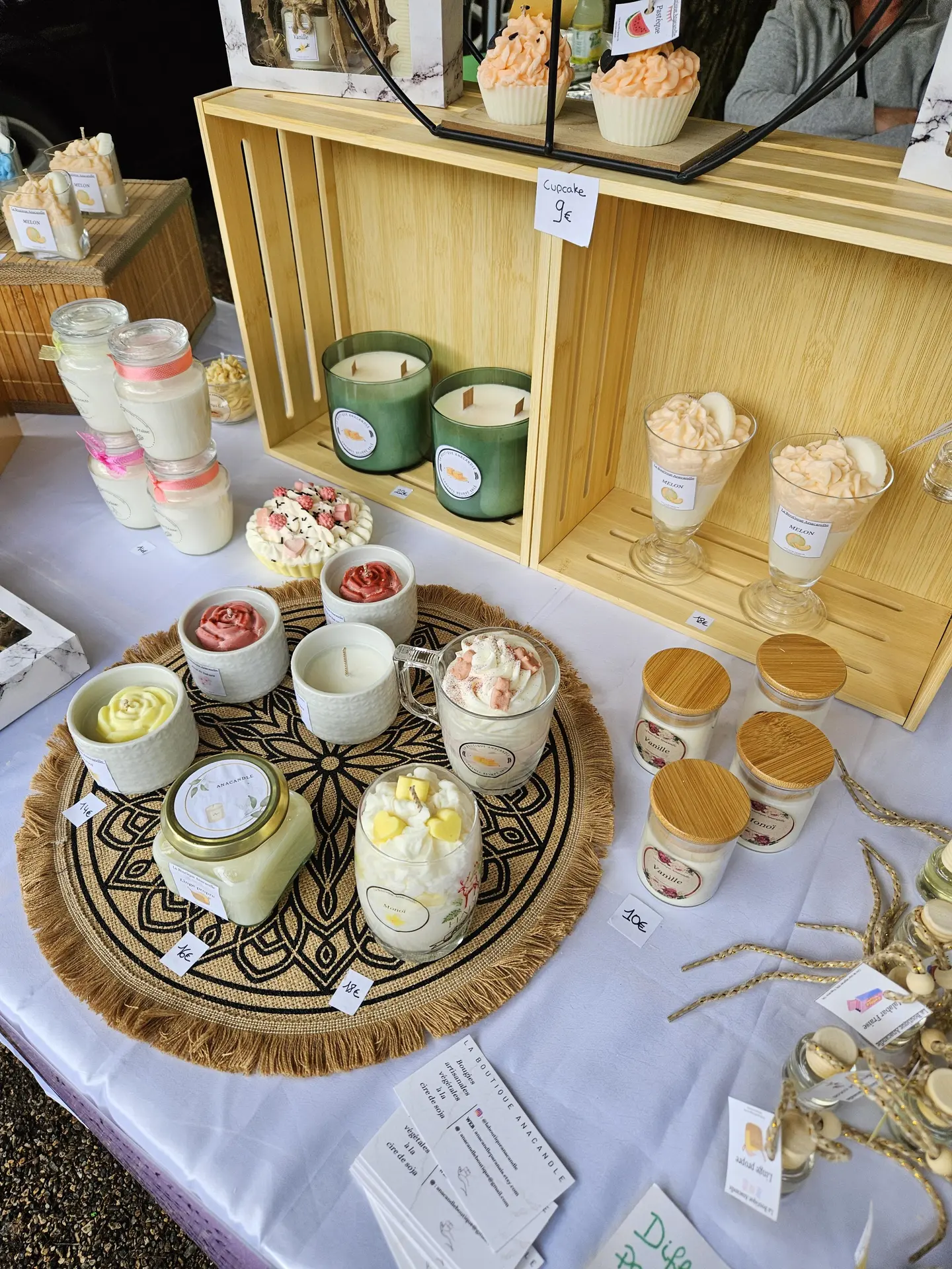 Ce stand de marché présente une variété de bougies artisanales en trompe-l'œil, de différentes tailles, formes et couleurs, disposées sur une table pour la vente.