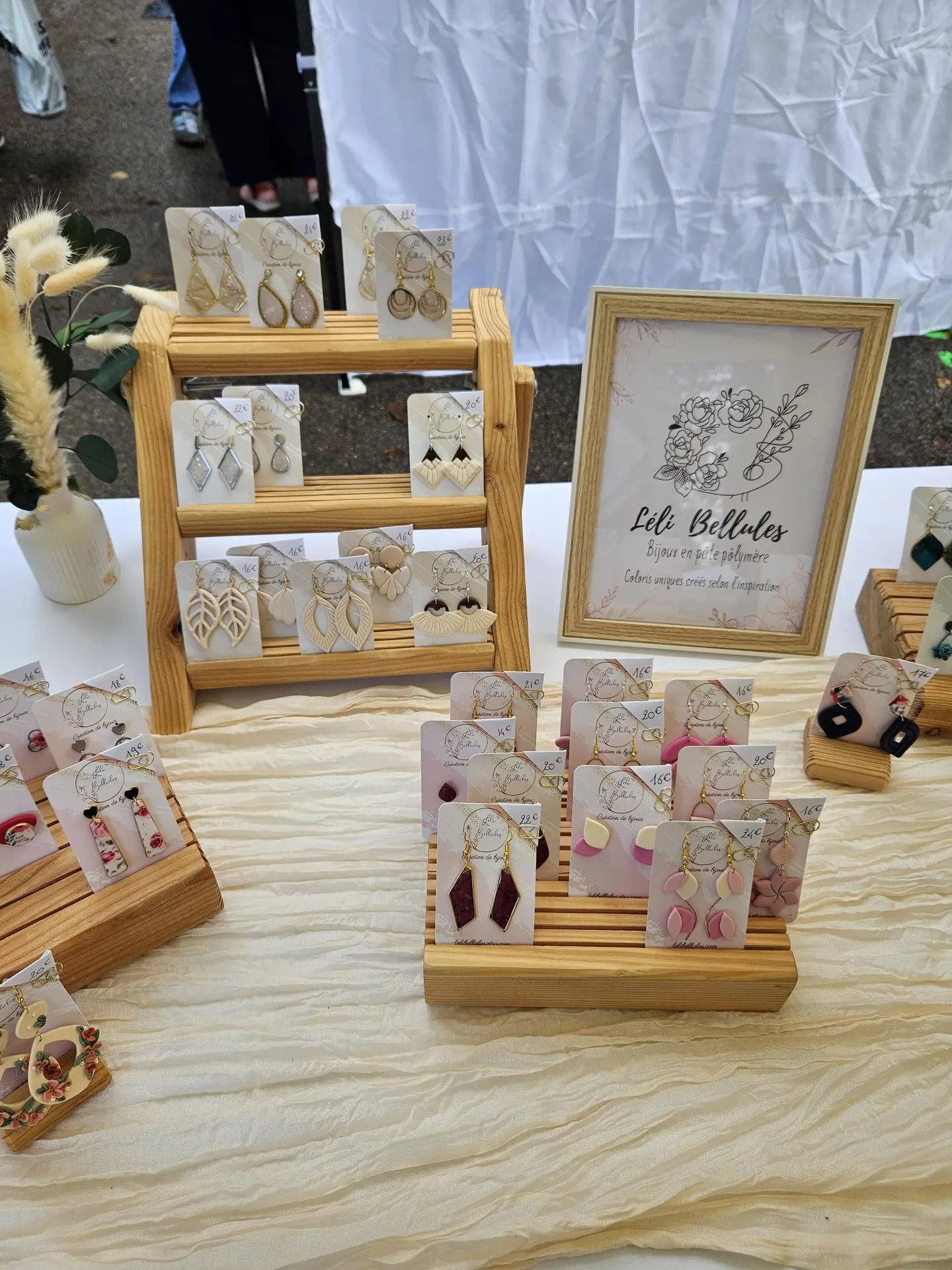 Ce stand de marché présente une variété de boucles d'oreilles artisanales, de différentes tailles, formes et matériaux, disposées sur des présentoirs en bois clair et sur une table en bois clair.