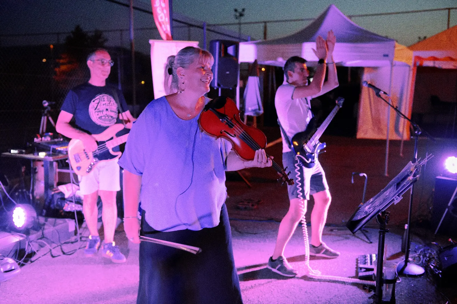 Une femme blonde tenant un violon se tient au centre, souriante, sur une scène extérieure éclairée par des lumières violettes et roses, avec deux hommes jouant de la guitare ou de la basse derrière elle.