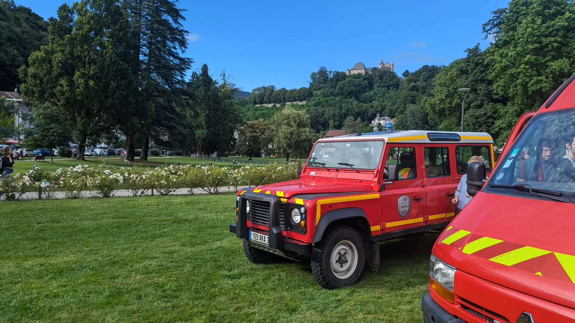 Sur une étendue d’herbe du parc d'Uriage, deux véhicules rouges des pompiers sont stationnés, entourés d’arbres et de massifs fleuris, avec en arrière-plan une colline boisée surmontée du château d'Uriage.