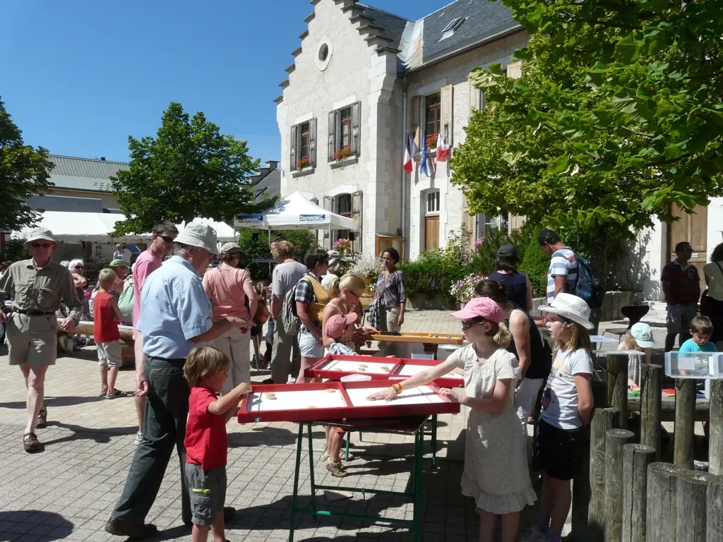 De nombreuses personnes regroupées sur la place devant la mairie et l'office de tourisme, dont des enfants au premier plan jouant à des jeux en bois sur une table