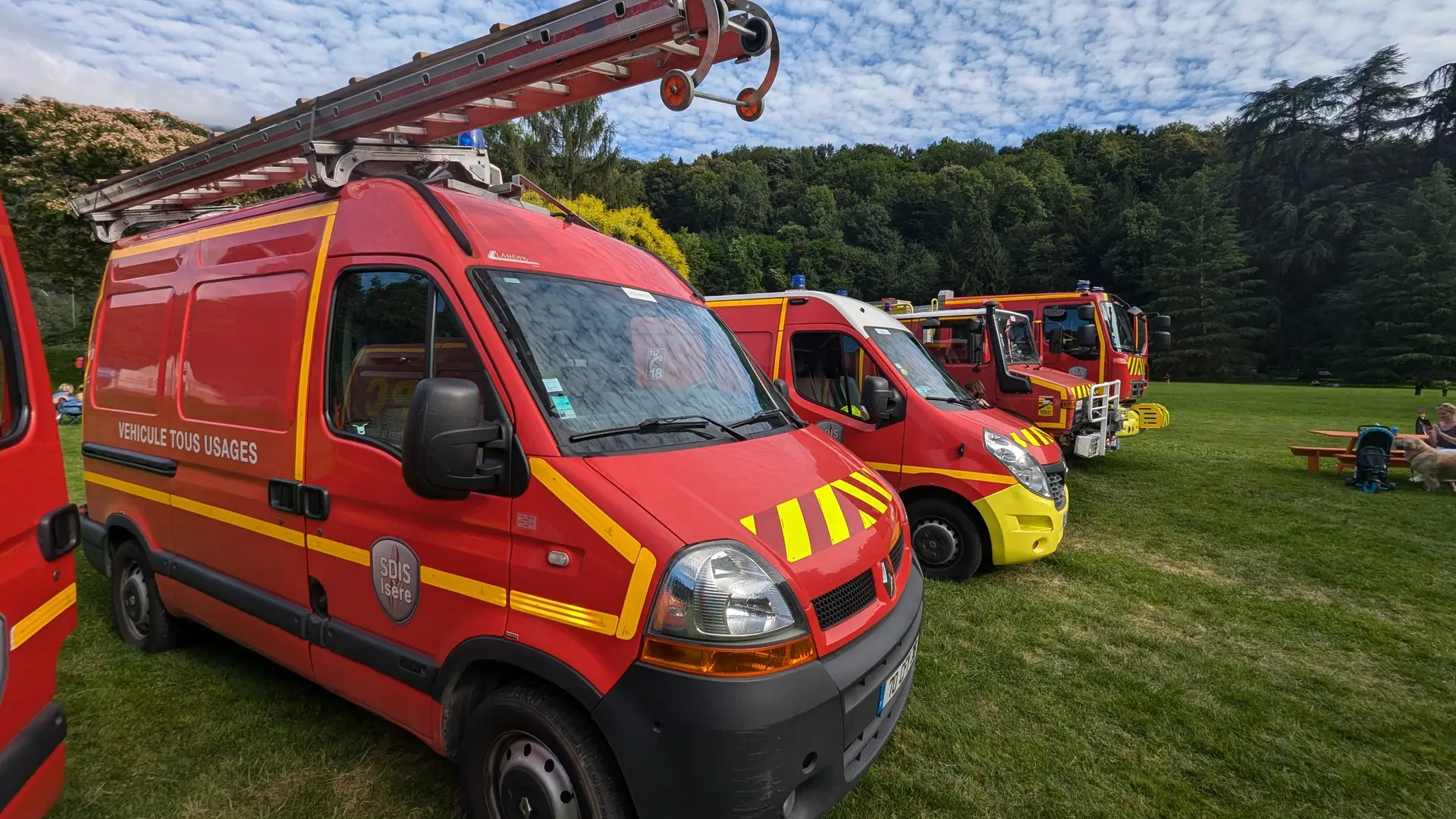 Plusieurs véhicules rouges des pompiers, alignés sur une pelouse du parc d’Uriage, sont exposés sous un ciel légèrement nuageux, avec quelques visiteurs et arbres en arrière-plan.