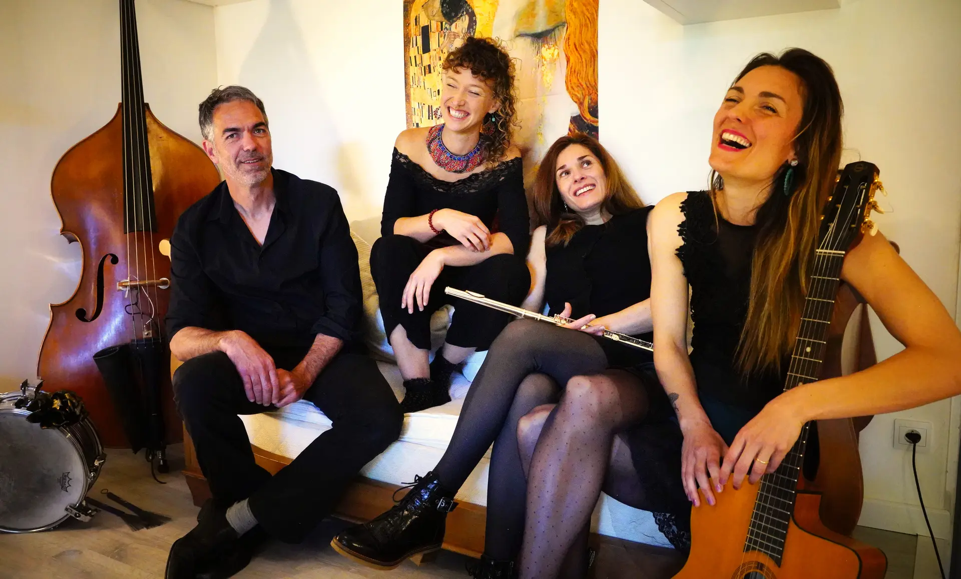 Un groupe de quatre musiciens assis, avec un homme à gauche près d'une contrebasse et d'une batterie, et trois femmes souriantes, dont une au centre tenant une flûte traversière et une autre à droite avec une guitare.