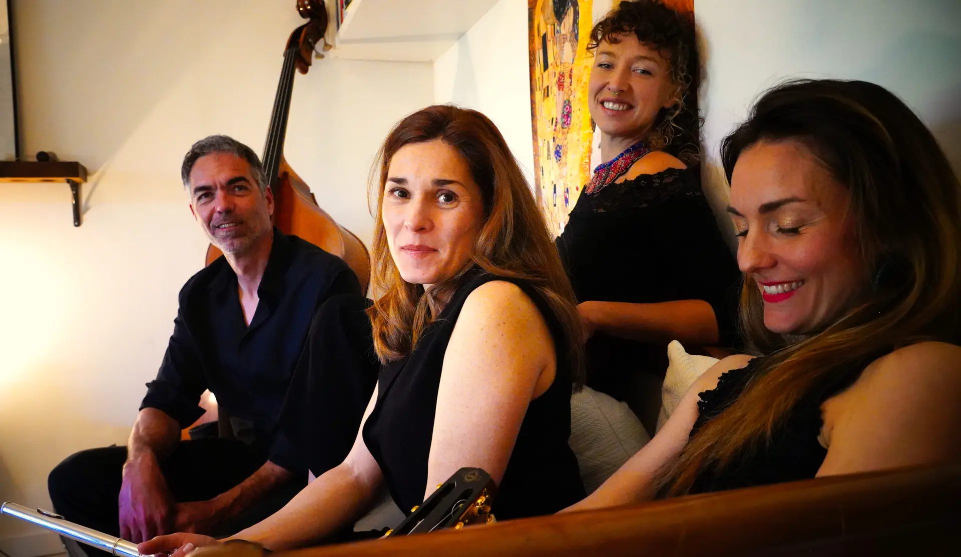 The Memory Box, un quatuor musical, avec un homme assis à gauche, une femme, une autre femme derrière elle souriant, et une quatrième femme à droite regardant vers le bas et souriant également, dans une ambiance intérieure chaleureuse.