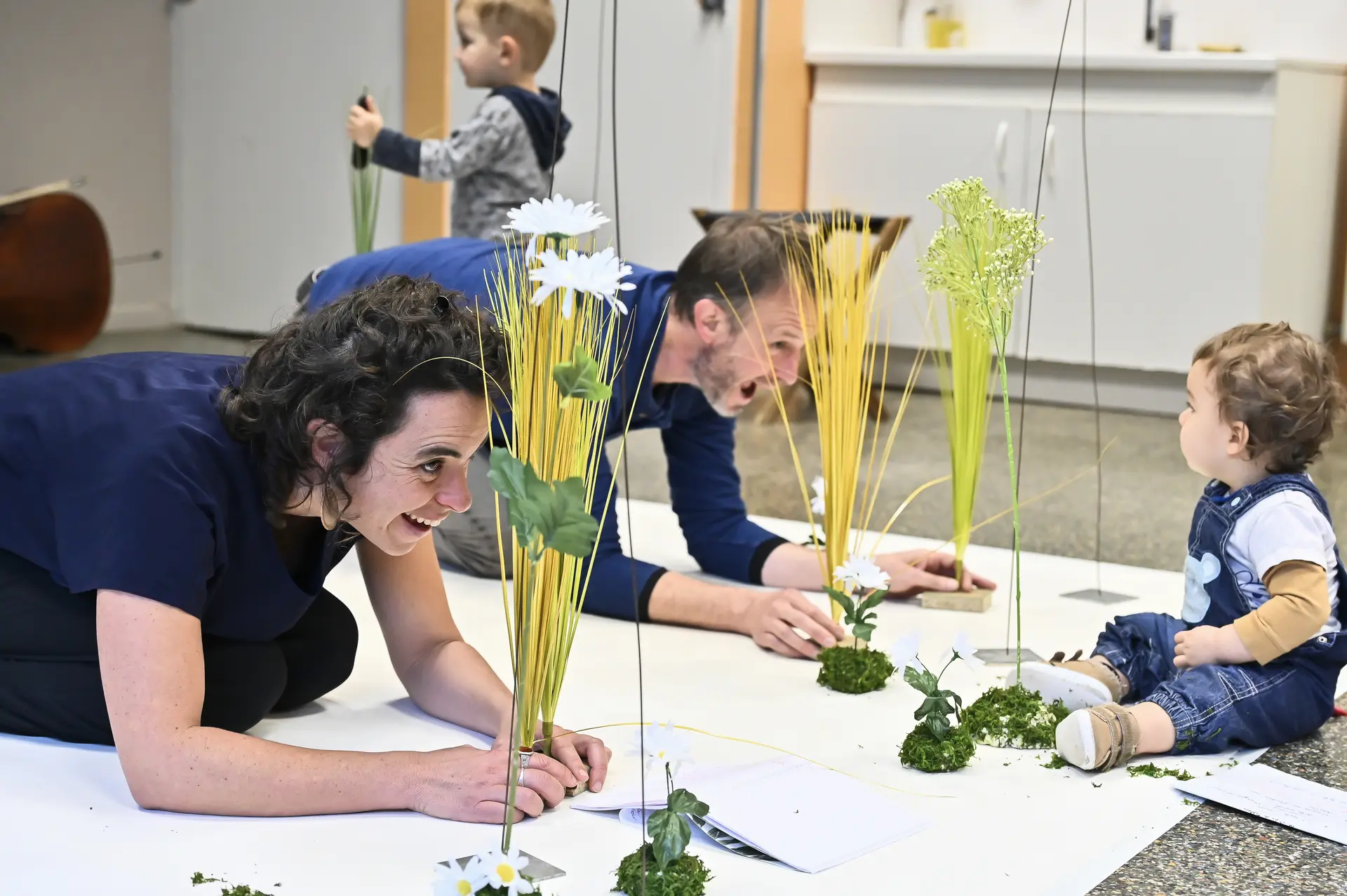 Photo représentant les comédiens en scène se cachant derrière des plantes artificielles devant un enfant en bas âge.