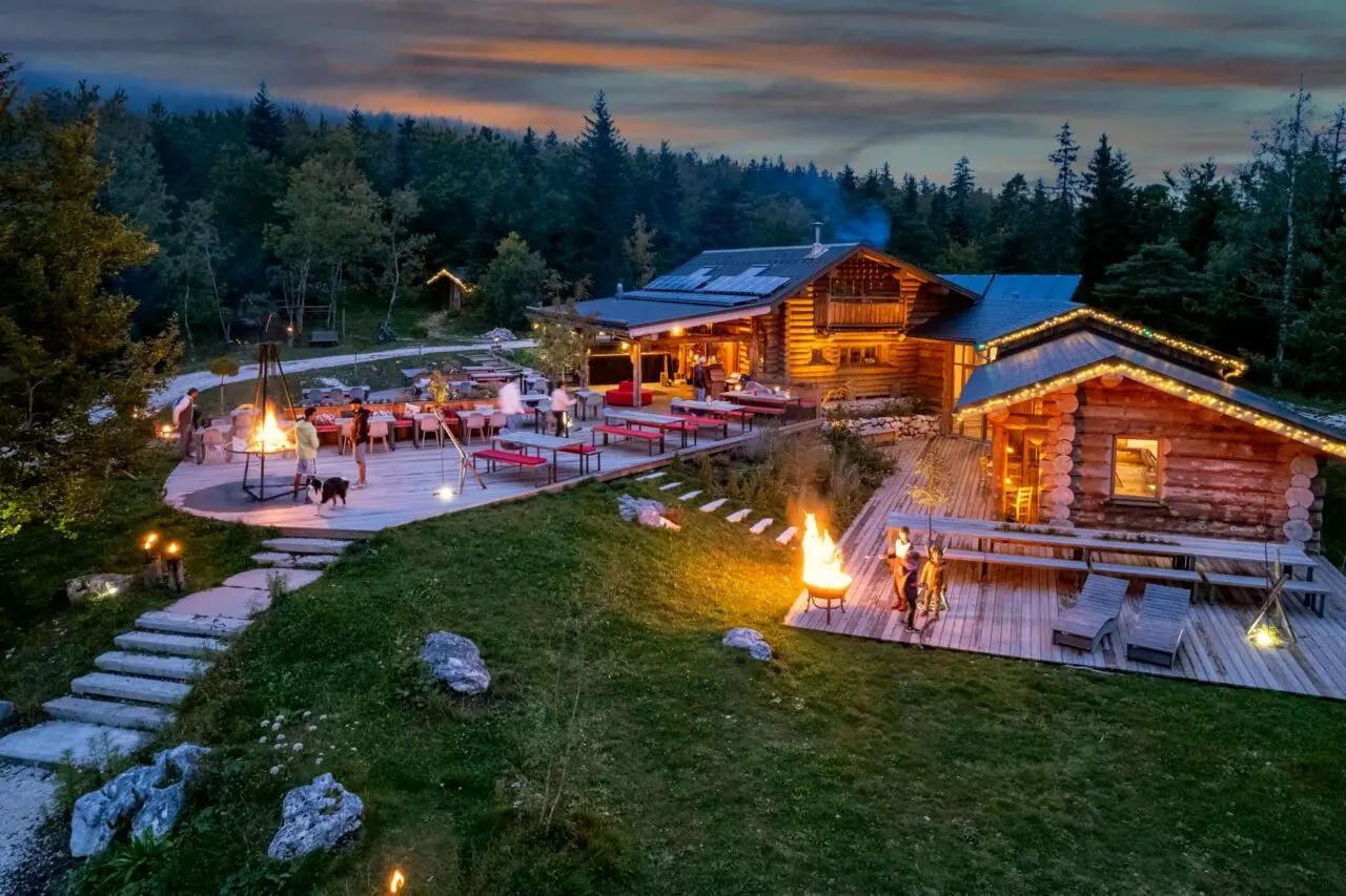 Photo d'un bâtiment ossature bois avec des guirlandes lumineuses au bord des toits. 2 terrasses en bois avec des tables, chaises et quelques transats, entourées d'espaces herbacés et d'une forêt de sapins : le restaurant du Clariant et ses terrasses.