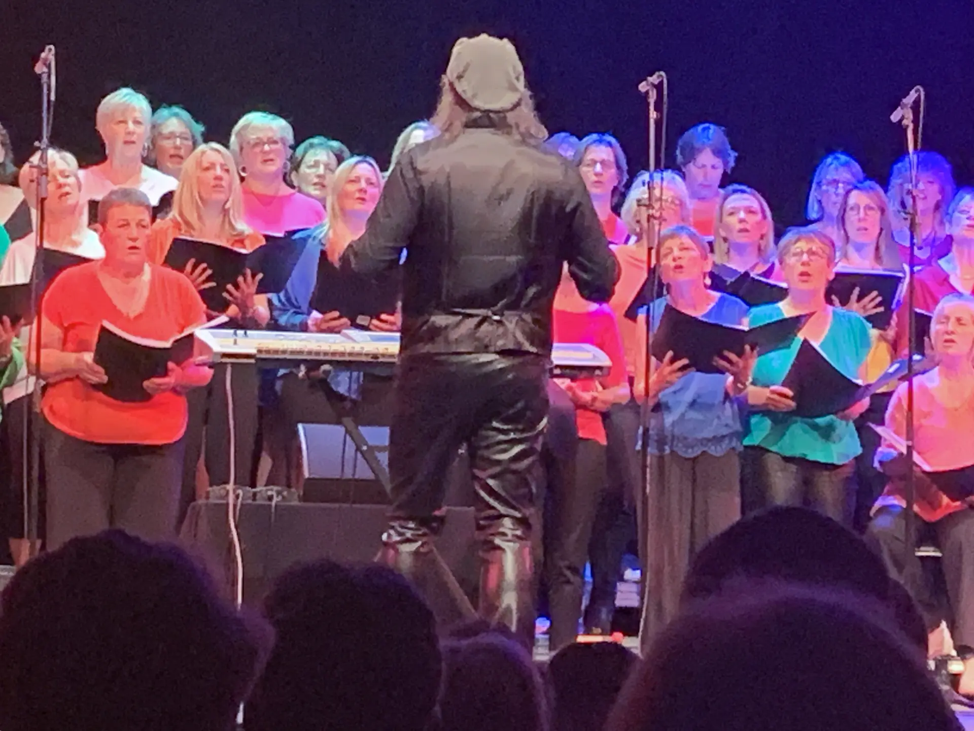 Un groupe de personnes chante, debout en rangs, visage éclairés par des lumières colorées. Au premier plan, le chef de coeur est vue de dos. La scène semble animée, avec des gestes expressifs des choristes