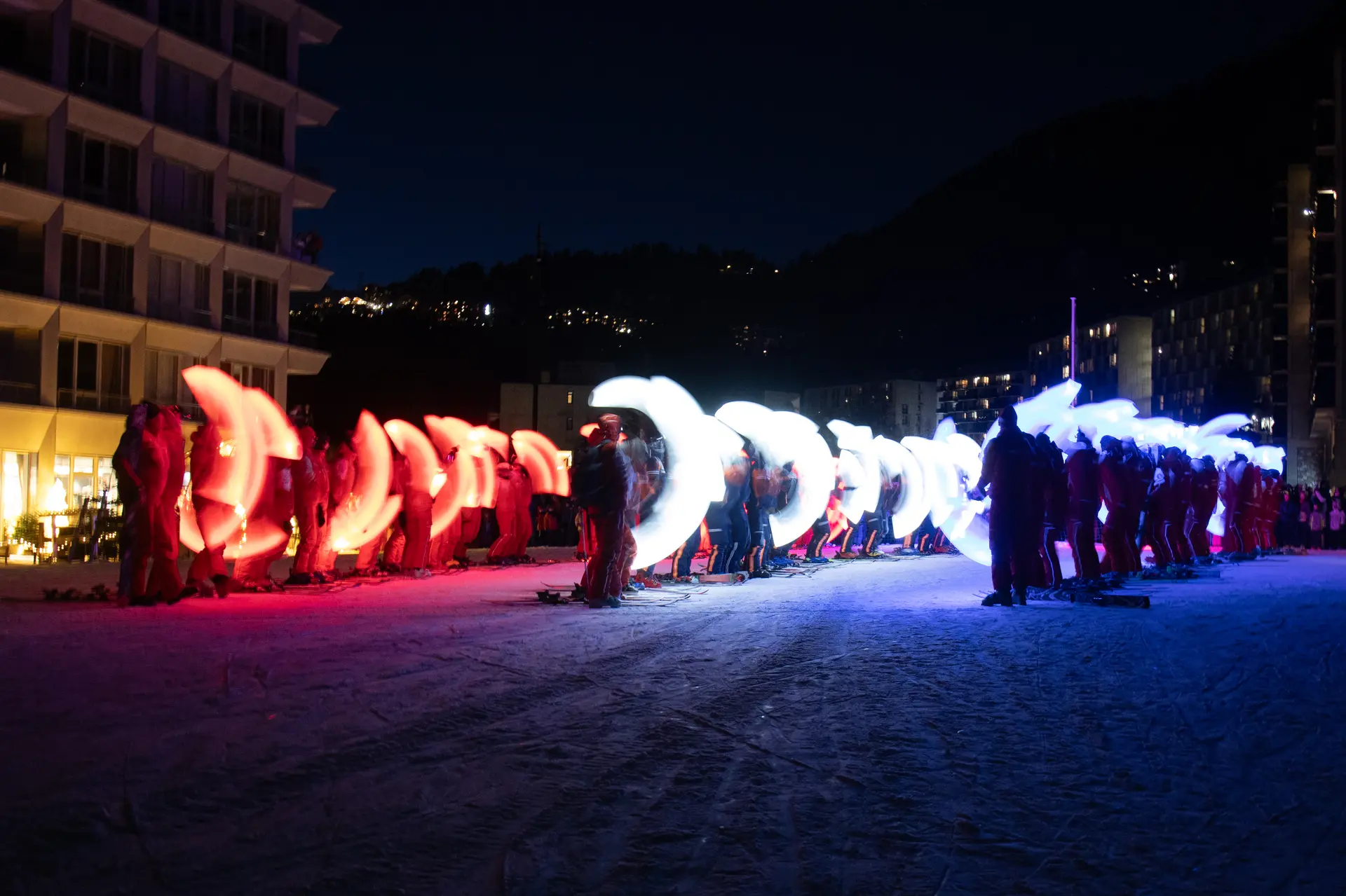 Arrivée de la descente sur le front de neige de Flaine. Les moniteurs ESF sont alignés et créent un drapeau tricolore avec leurs bâtons lumineux. La neige est éclairée successivement de rouge, de blanc et de bleu, encadrée par les façades des immeubles dans la nuit.