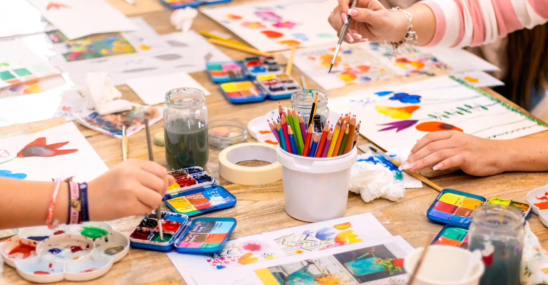 Zoom sur une table recouverte d'ustencils créatifs, mains d'enfant et d'adulte en train de peindre