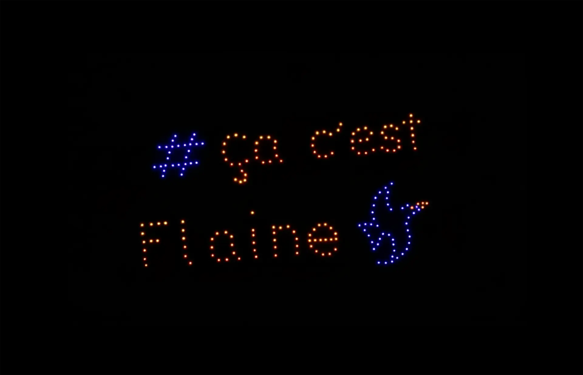 Spectacle nocturne de drones sur fond noir. Des points lumineux forment le texte orange « # ça c’est Flaine », avec à droite le logo bleu de la station de ski, un choucas.