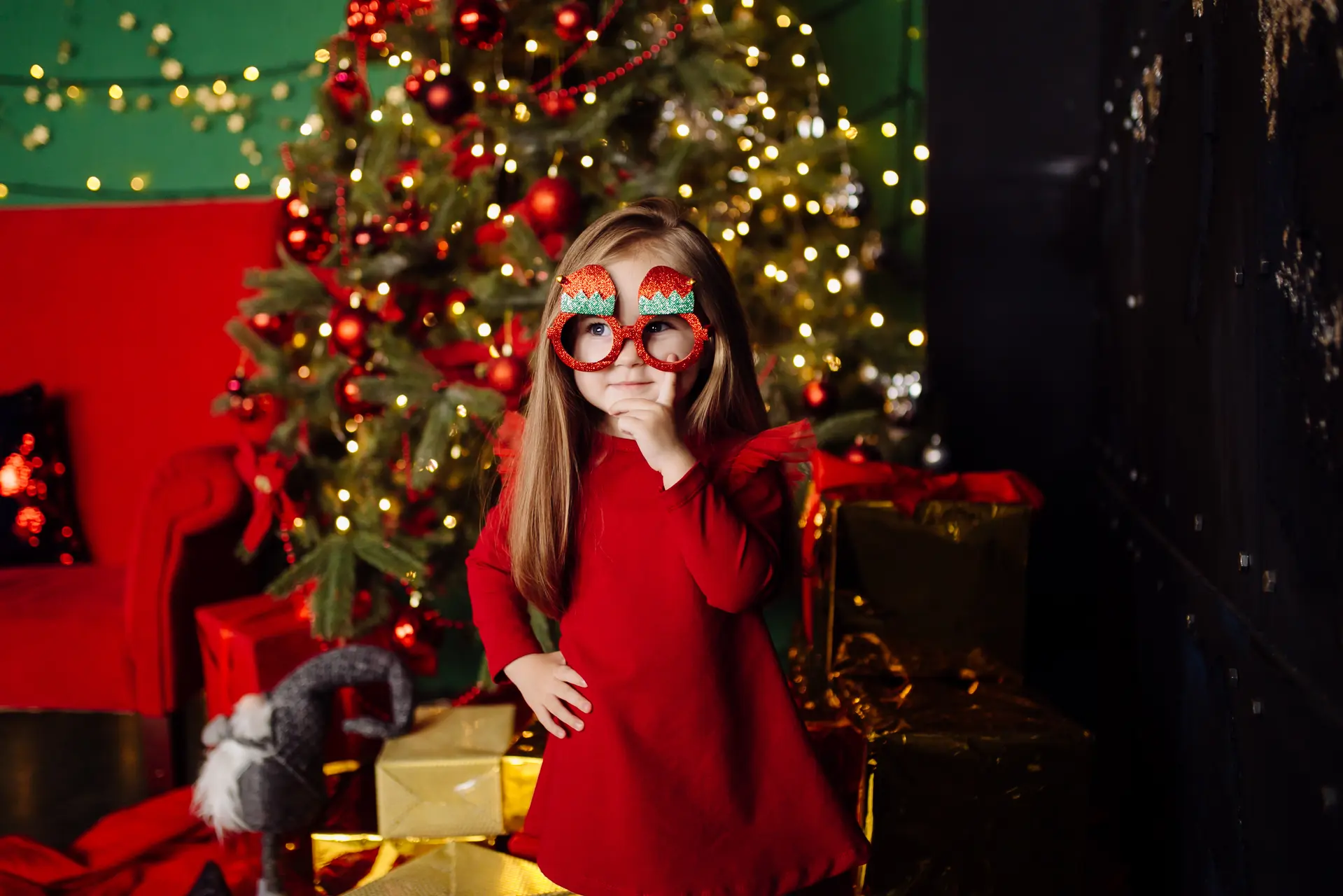 une petite fille avec une robe rouge et des lunettes rouge, se tient devant un sapin de Noël