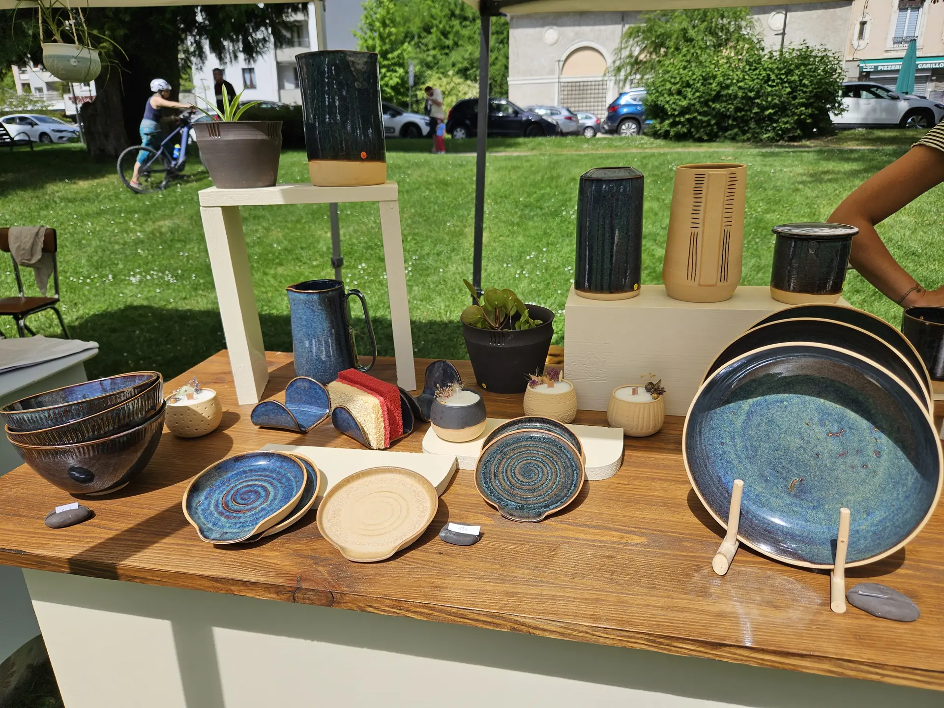 Un stand du marché des potiers en plein air présentant diverses poteries artisanales, principalement des assiettes, des bols et des vases aux émaux bleu profond et terre cuite, disposés sur une table en bois et des supports blancs sur un fond de parc ensoleillé.