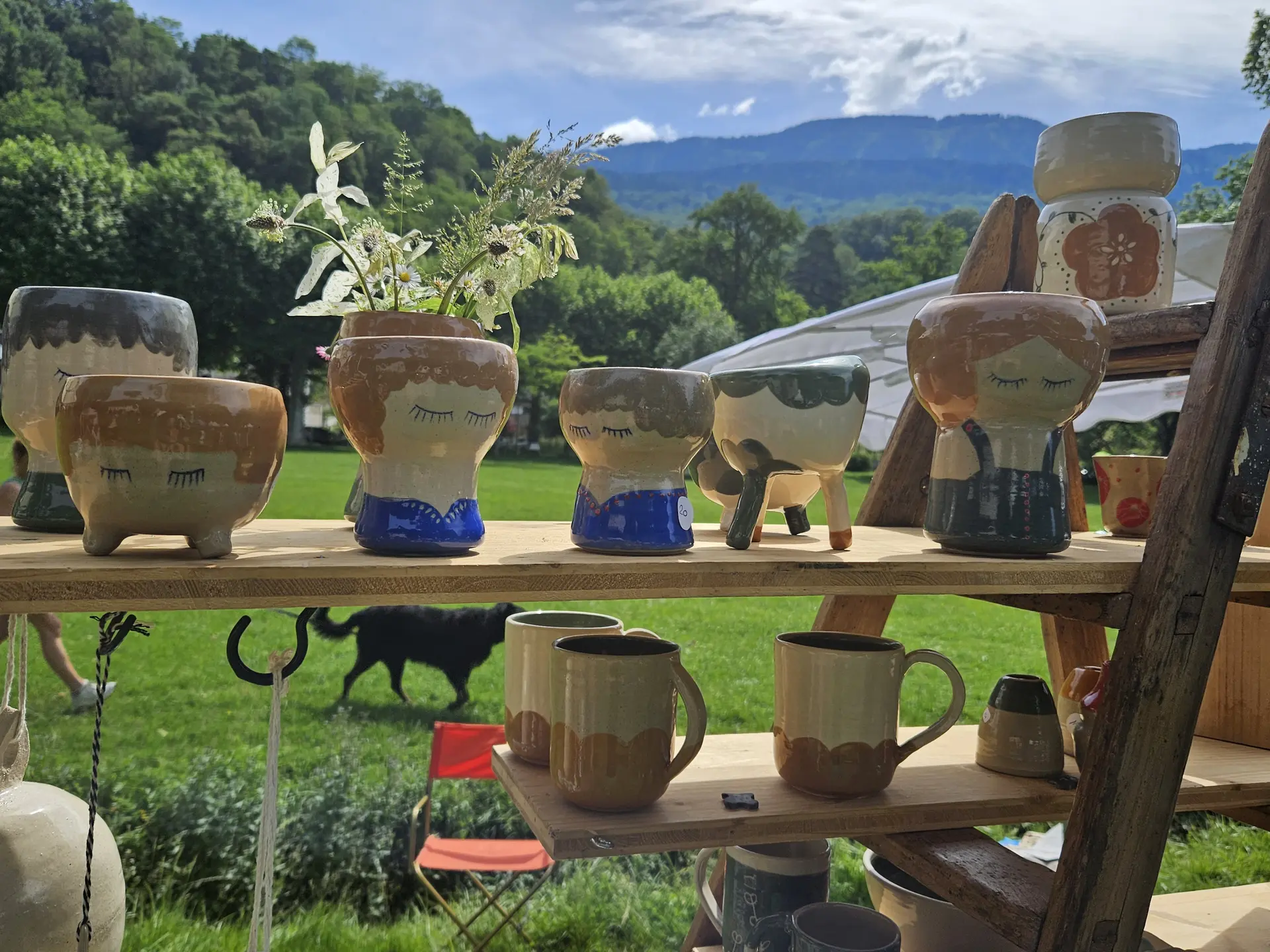 Un étalage en bois sur deux niveaux présentant des poteries illustrées de visages stylisés et des mugs aux tons ocre, avec en arrière-plan un paysage verdoyant de montagnes