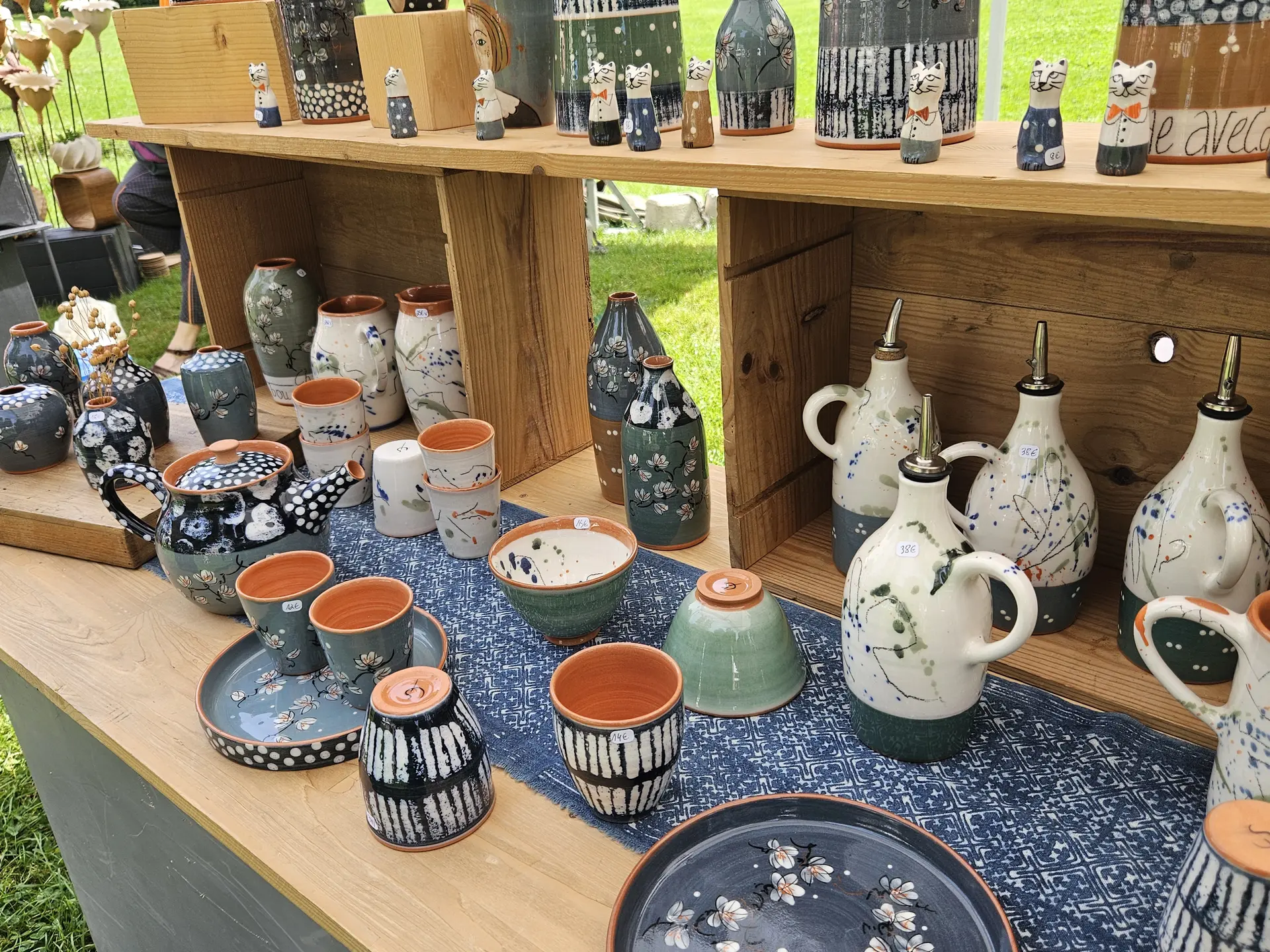 Un étalage en bois sur deux niveaux richement décoré de céramiques artisanales aux motifs printaniers, incluant des théières, des bols et des bouteilles d'huile ornés de fleurs de cerisier, ainsi que de petites figurines de chats stylisées disposées sur l'étagère supérieure.