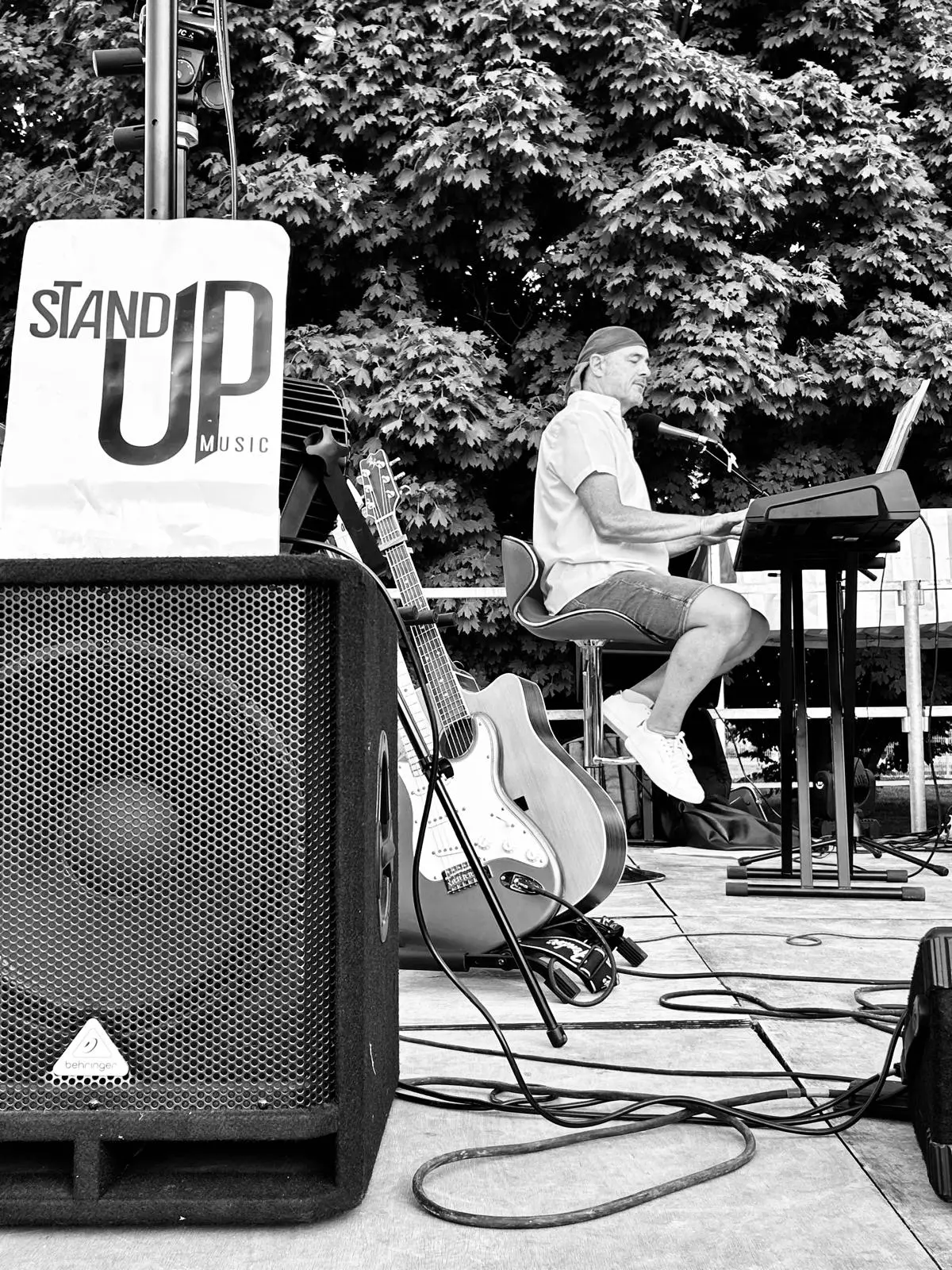 Un homme est assis et joue d'un instrument ou utilise un ordinateur portable en arrière-plan, tandis qu'au premier plan, à gauche, se trouve une grande enceinte audio et un panneau affichant les mots « STAND UP MUSIC », avec un instrument à cordes posé à côté et des arbres derrière l'homme.