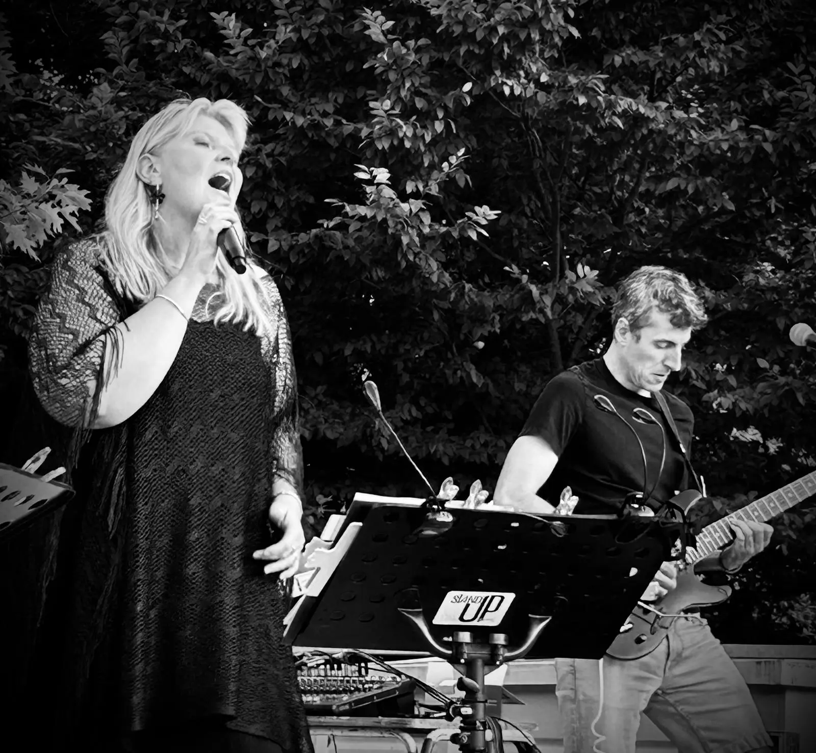 Une femme blonde chante dans un microphone tout en se tenant devant un pupitre à musique, avec un homme jouant d'un instrument ou utilisant de l'équipement musical à sa droite et des arbres en arrière-plan.