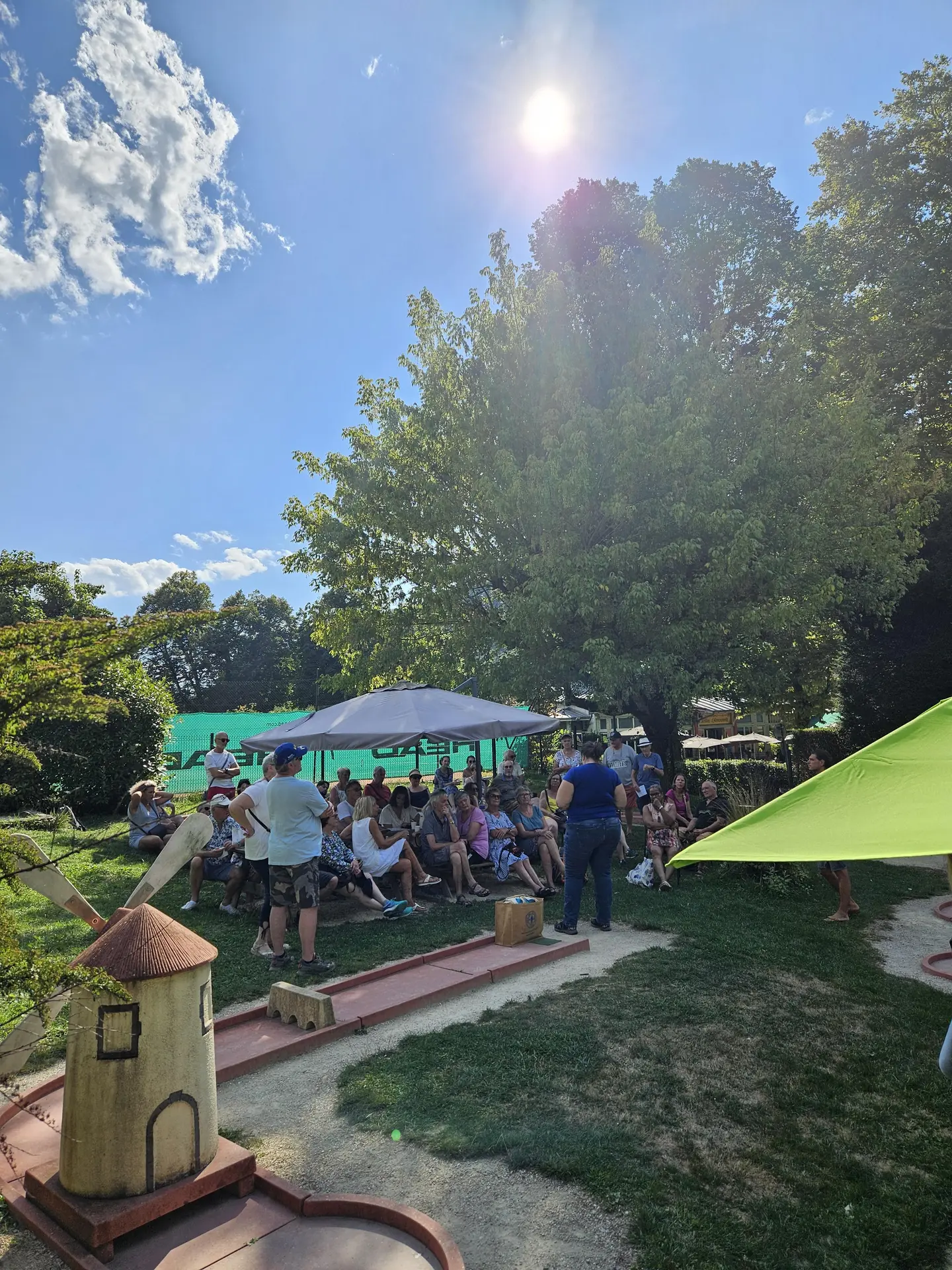 Une foule de personnes est rassemblée sous un grand arbre et des parasols sur un terrain de mini-golf ensoleillé, entouré de verdure.