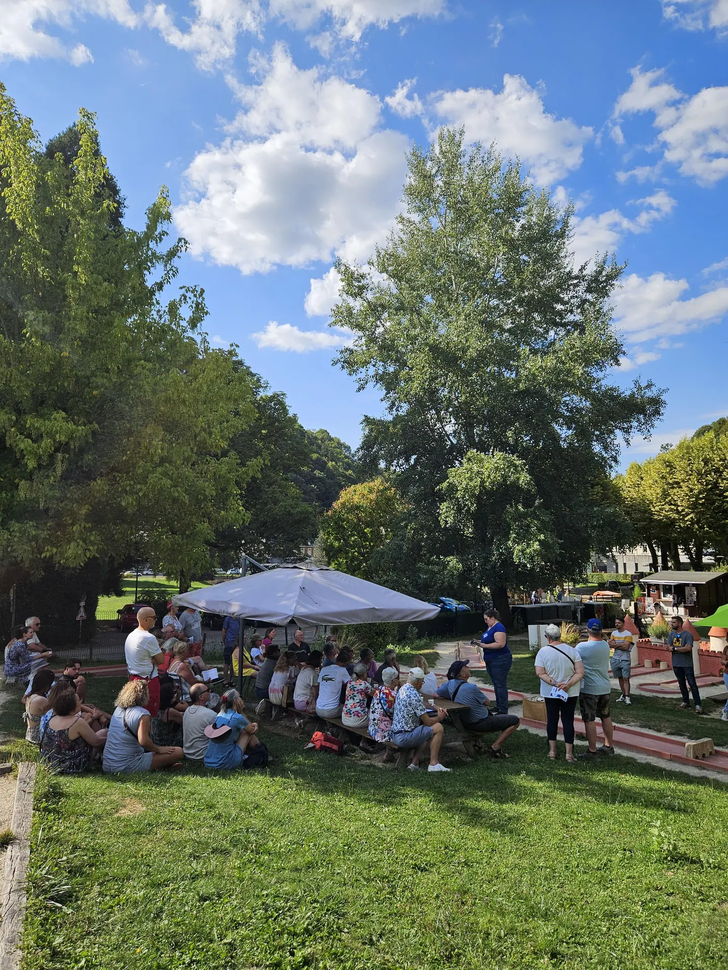 Une foule de personnes est rassemblée sous un grand arbre et des parasols sur un terrain de mini-golf ensoleillé, entouré de verdure.