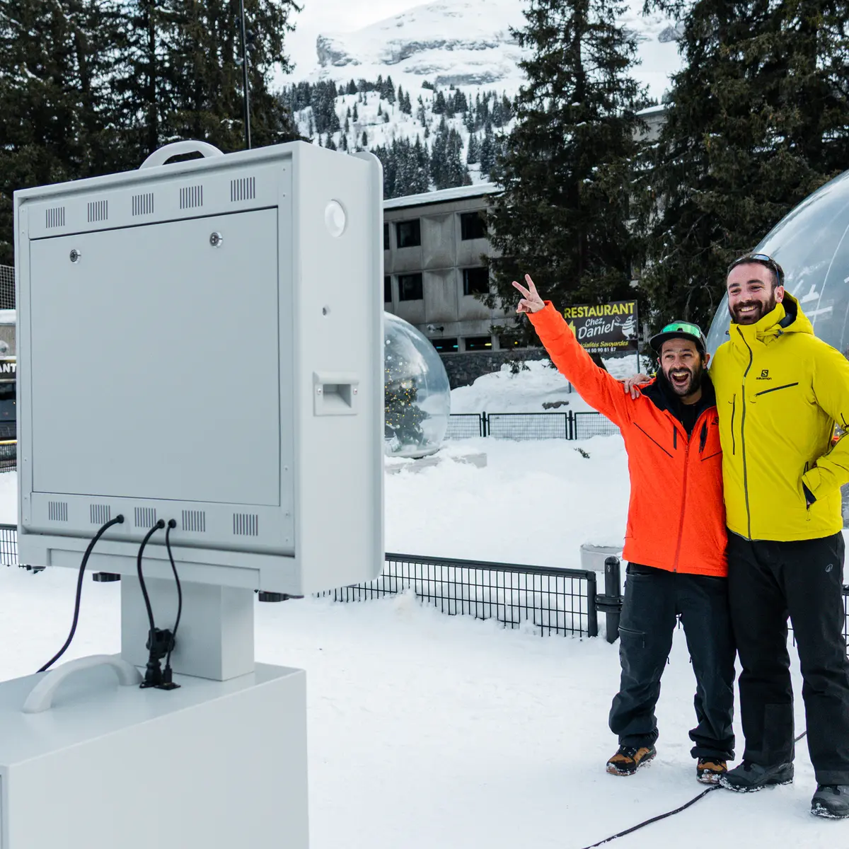 Deux hommes en tenue de ski sourient et posent devant une borne photo installée dans une station de montagne enneigée.