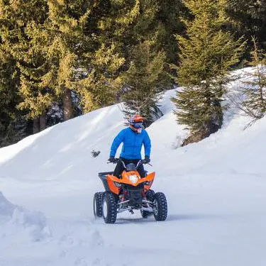 Une personne portant une veste bleue et un casque orange conduit un quad orange et noir sur un chemin enneigé.
