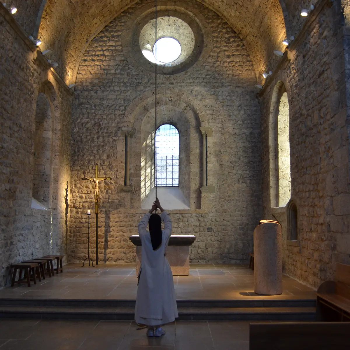 Au coeur de l'édifice éclairé d'une lumière filtrée par les vitraux, une personne en habit religieux s'apprête à tirer la ficelle reliée à la cloche de l'Église