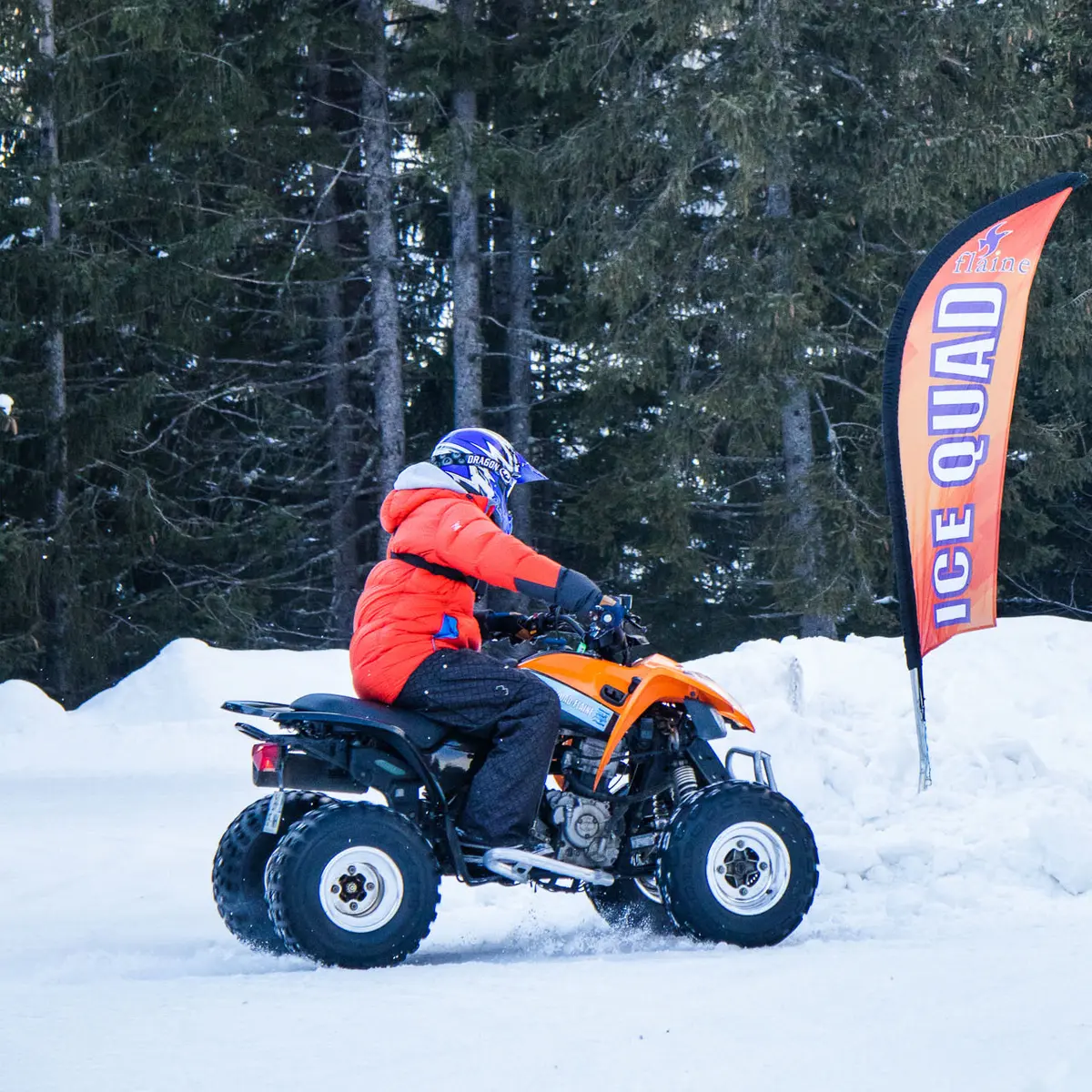 Une personne en veste orange et casque bleu conduit un quad noir et orange de profil. Le véhicule roule sur une piste enneigée devant une forêt de sapins.