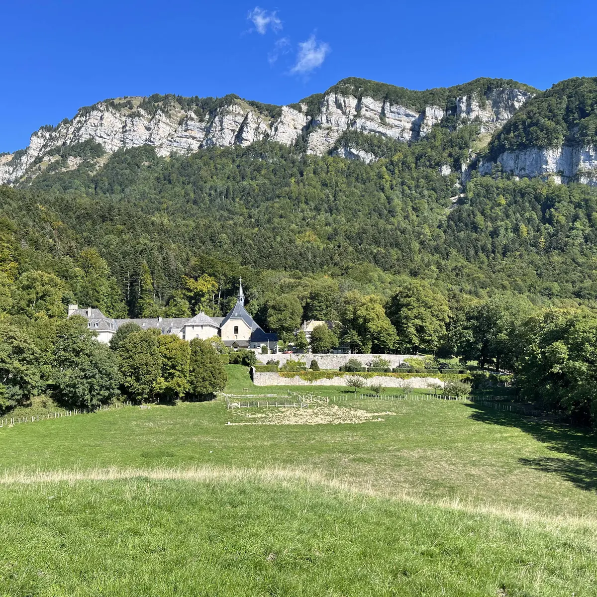 Splendide vue du Monastère, fondu dans son environnement verdoyant où la nature est omniprésente. Au premier plan, une prairie amène le regard jusqu'à l'édifice religieux, au pied de la montagne sous ciel bleu