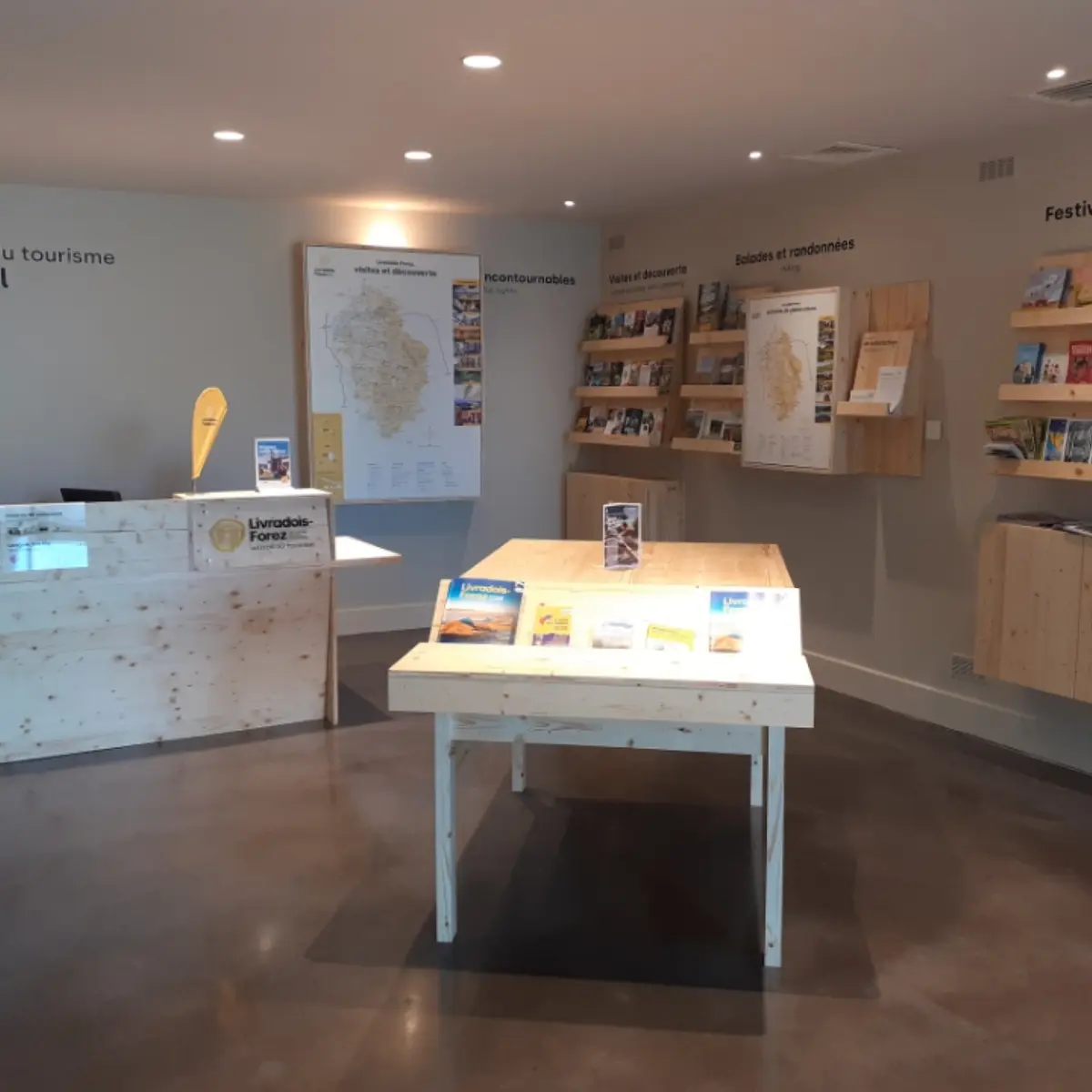 Intérieur du Point d'information touristique avec meubles de présentation pour la documentation du territoire. Au centre se trouve le piano pour échanger avec le conseiller en séjour. L'espace est ouvert et accueillant avec du mobilier en bois de la région Livradois-Forez