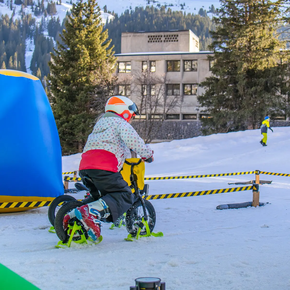 Un enfant portant un casque et une veste colorée s'entraîne sur un petit vélo-ski à l'intérieur d'un parcours balisé sur la neige.