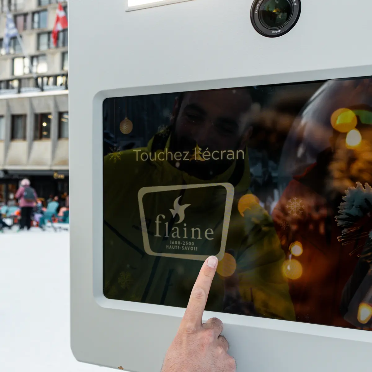 Une personne vêtue d'une veste jaune touche l'écran d'un photobooth installé en extérieur dans la neige.
