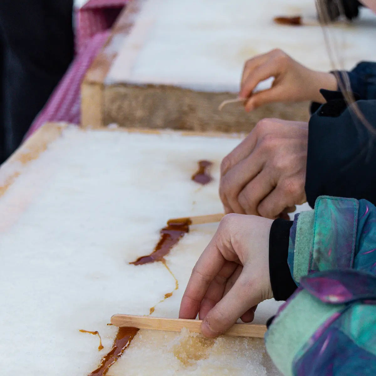 Des mains enroulent du sirop d'érable chaud sur des bâtonnets en bois directement sur la neige. C'est une façon amusante de transformer le sirop en bonbons glacés.
