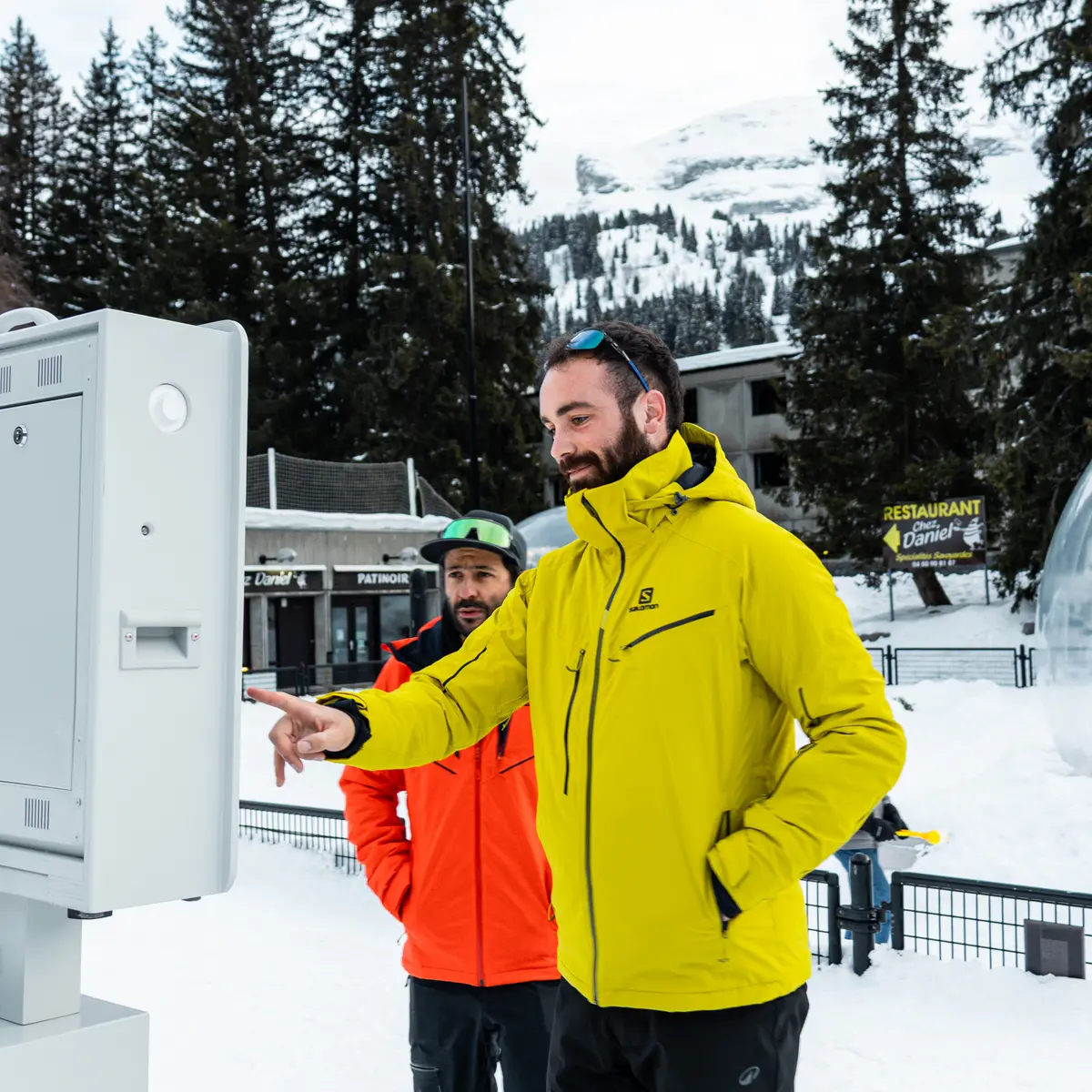 Deux hommes en tenue de ski utilisent une borne photo interactive dans une station enneigée, l'un pointant l'écran du doigt. Ils se trouvent en extérieur, entourés de sapins, d'une bulle décorative géante et de bâtiments de montagne.