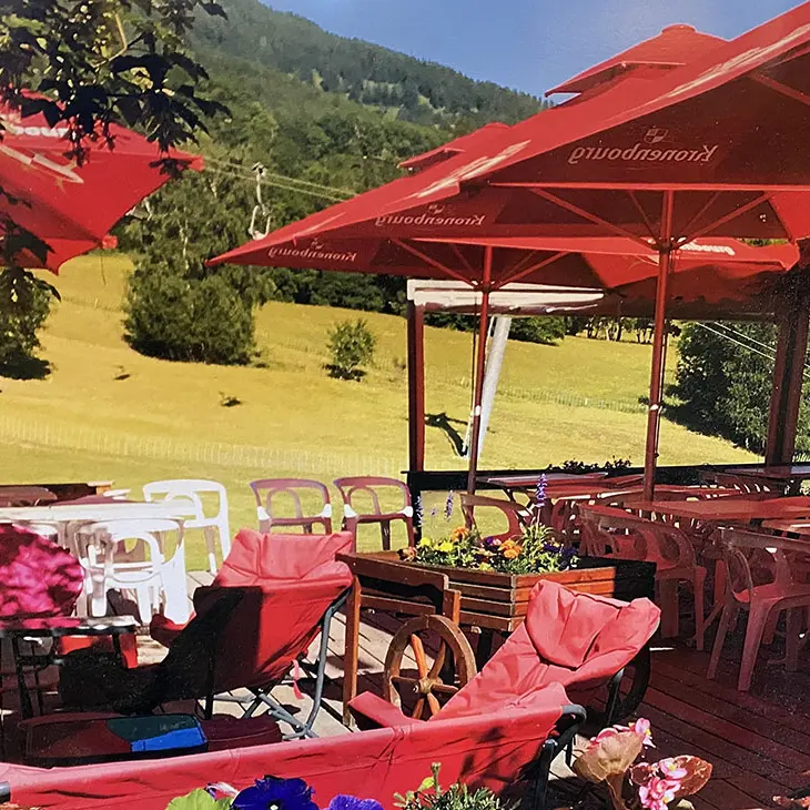 Terrasse extérieure du restaurant en été, tables et chaises en plastique, grands parasols, vue sur un champ et sur les montagnes