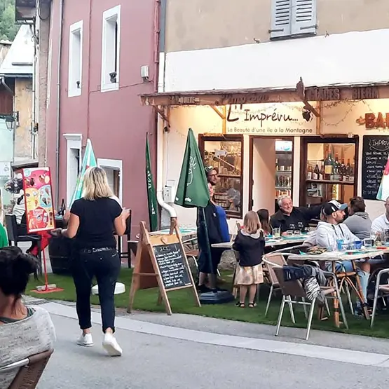 Terrasse extérieure du restaurant, située dans la grand rue d'Allos, fausse pelouse au sol, tables et chaises. Inscription sur façade : 'L'Imprévu, La Beauté d'une île à la montagne