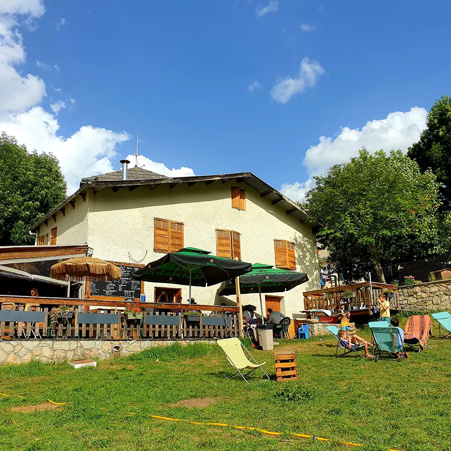 Overview of the restaurant, a two-story stone and wood building with a wooden terrace on the ground floor, tables, chairs, parasols, and sun loungers. Meadow in front of the restaurant with sun loungers.